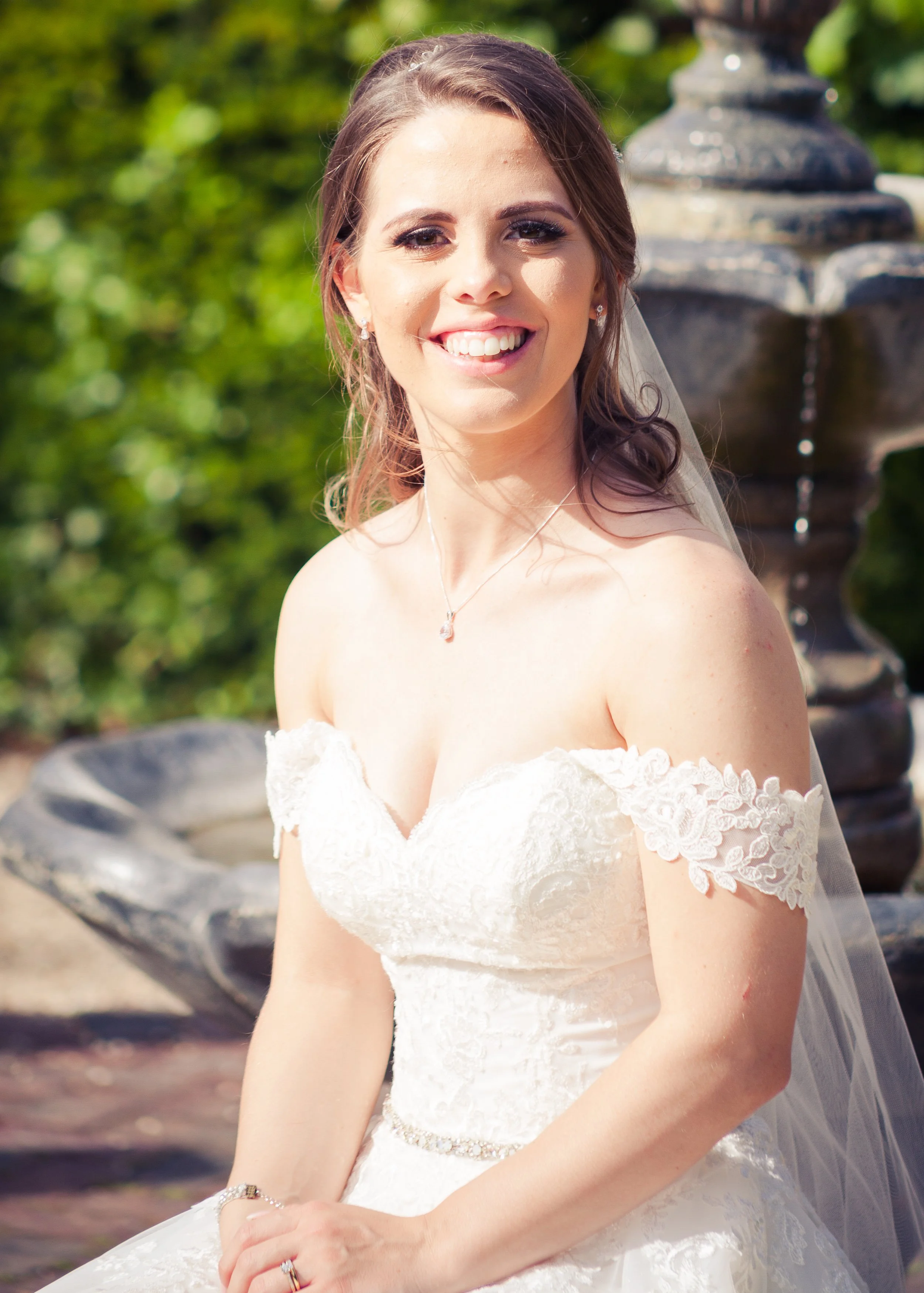 Close-up of a smiling bride in a white lace wedding dress, wearing a necklace and earrings, with a garden and fountain in the background.