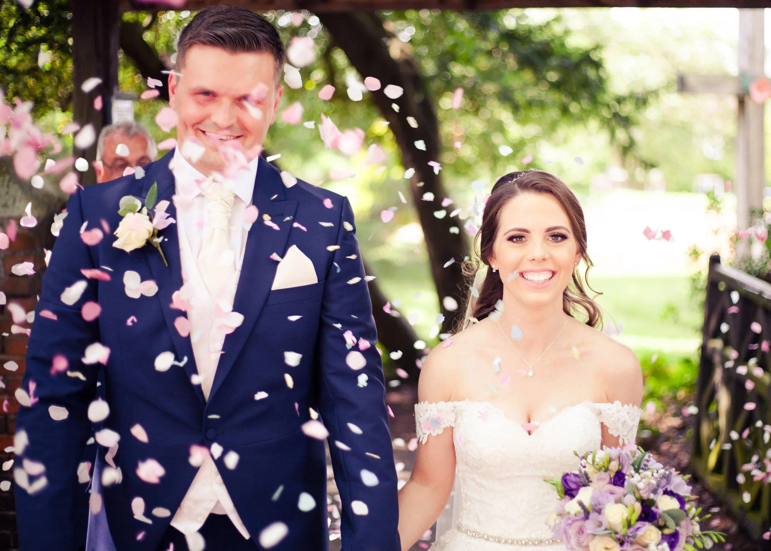 A smiling bride and groom at their outdoor wedding, surrounded by falling pink and white confetti.