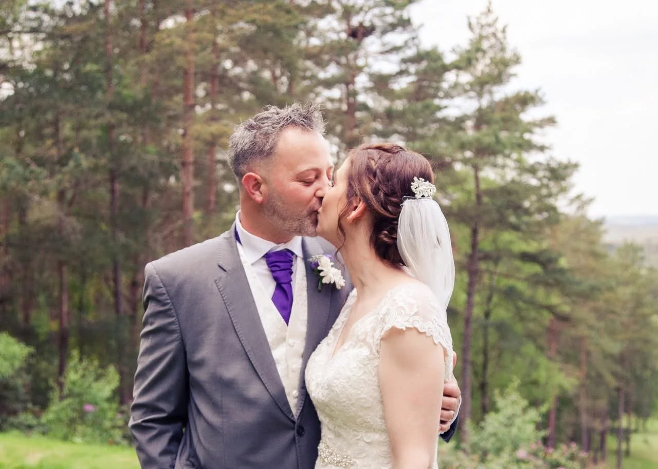 A bride and groom kissing outdoors in a forested area on their wedding day.