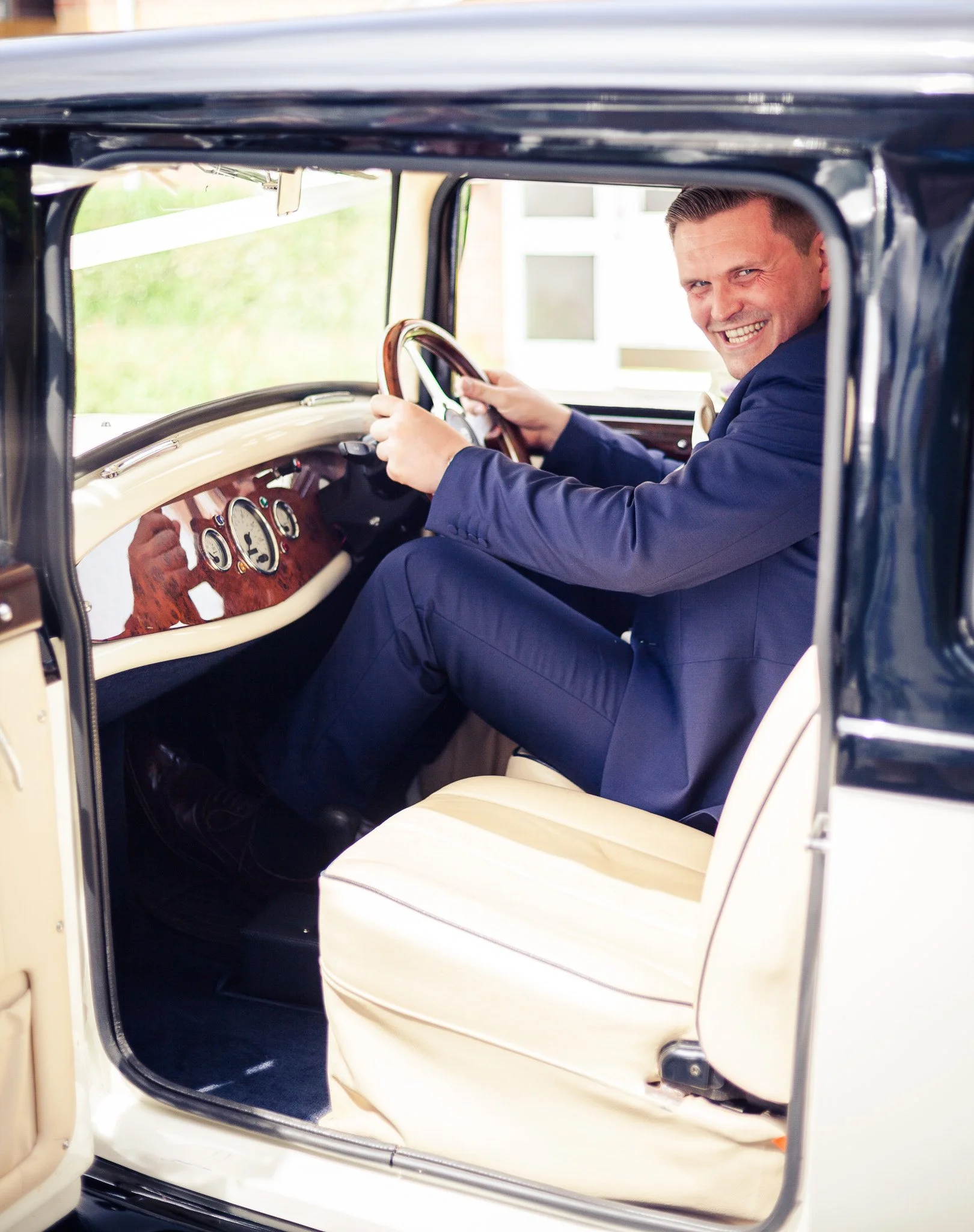 A man in a blue suit sitting in the driver's seat of a vintage cream-colored car, smiling and looking at the camera through the open window.