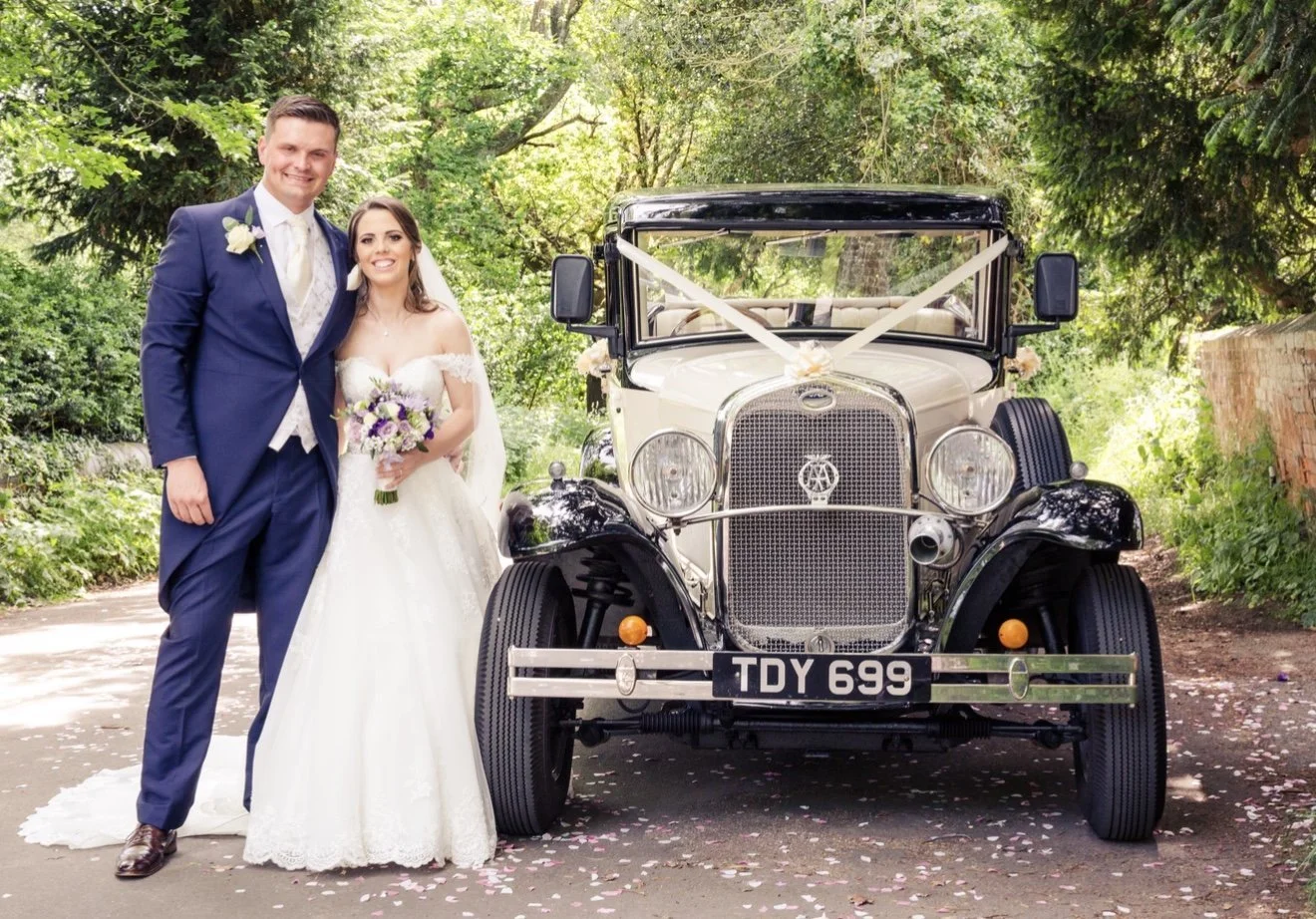 A bride and groom standing next to a vintage white wedding car decorated with ribbons, on a tree-lined path with fallen flower petals.