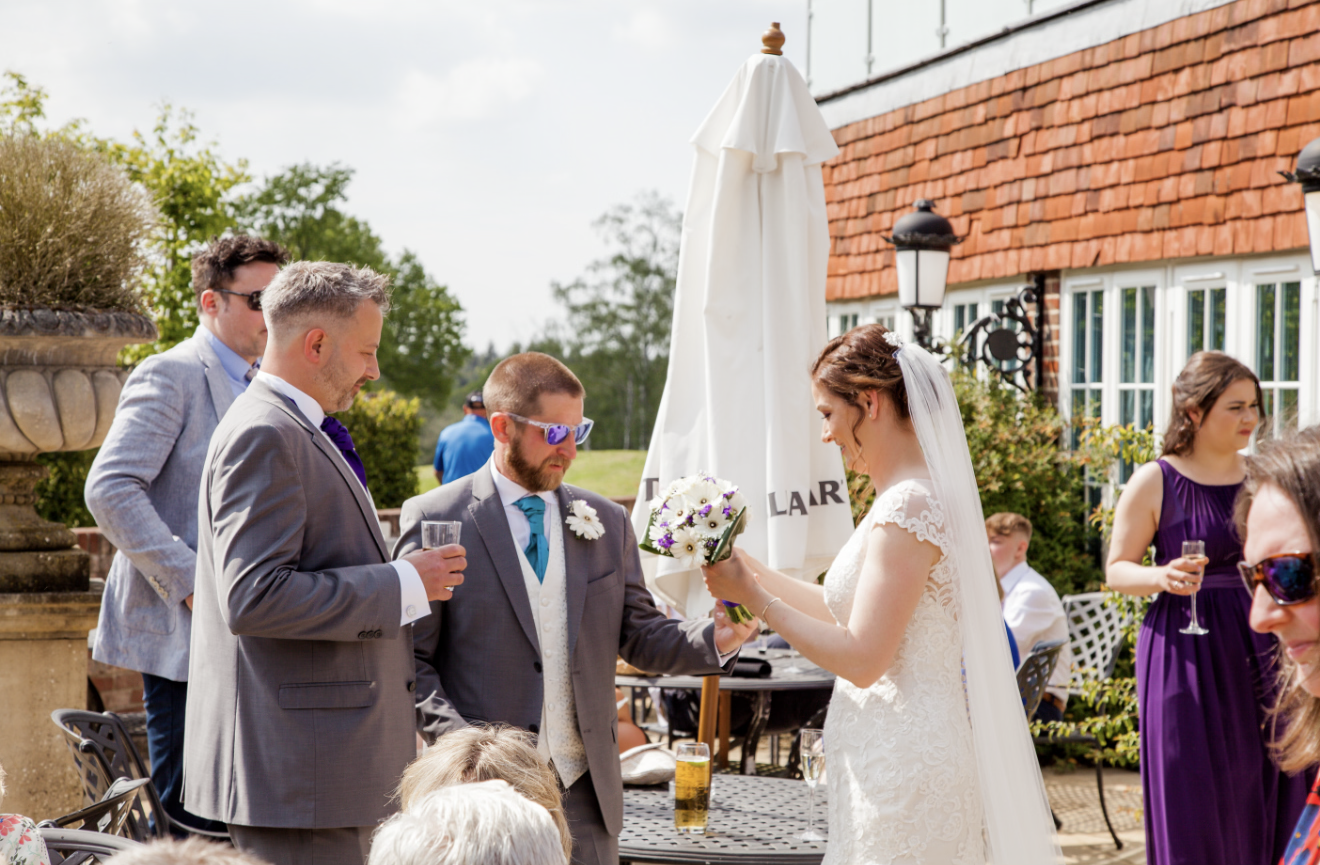 Bride in a white lace wedding gown holding a bouquet of flowers, receiving a toast or gift from a man in a gray suit at an outdoor wedding celebration, with other guests in formal attire and a building with a red tiled roof in the background.