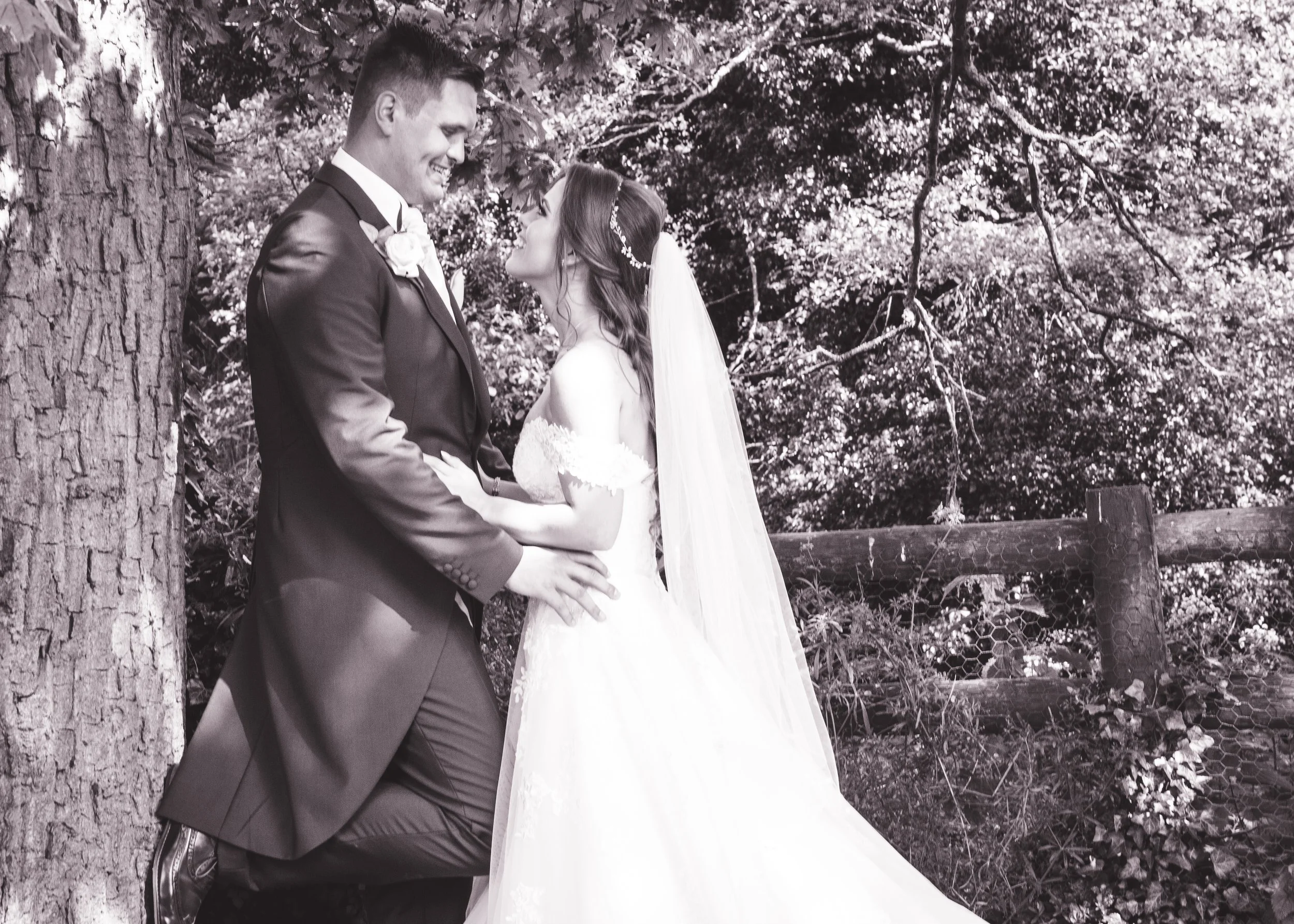 A black-and-white photo of a bride and groom standing near a large tree outdoors, gazing into each other's eyes and smiling, with trees and foliage in the background.