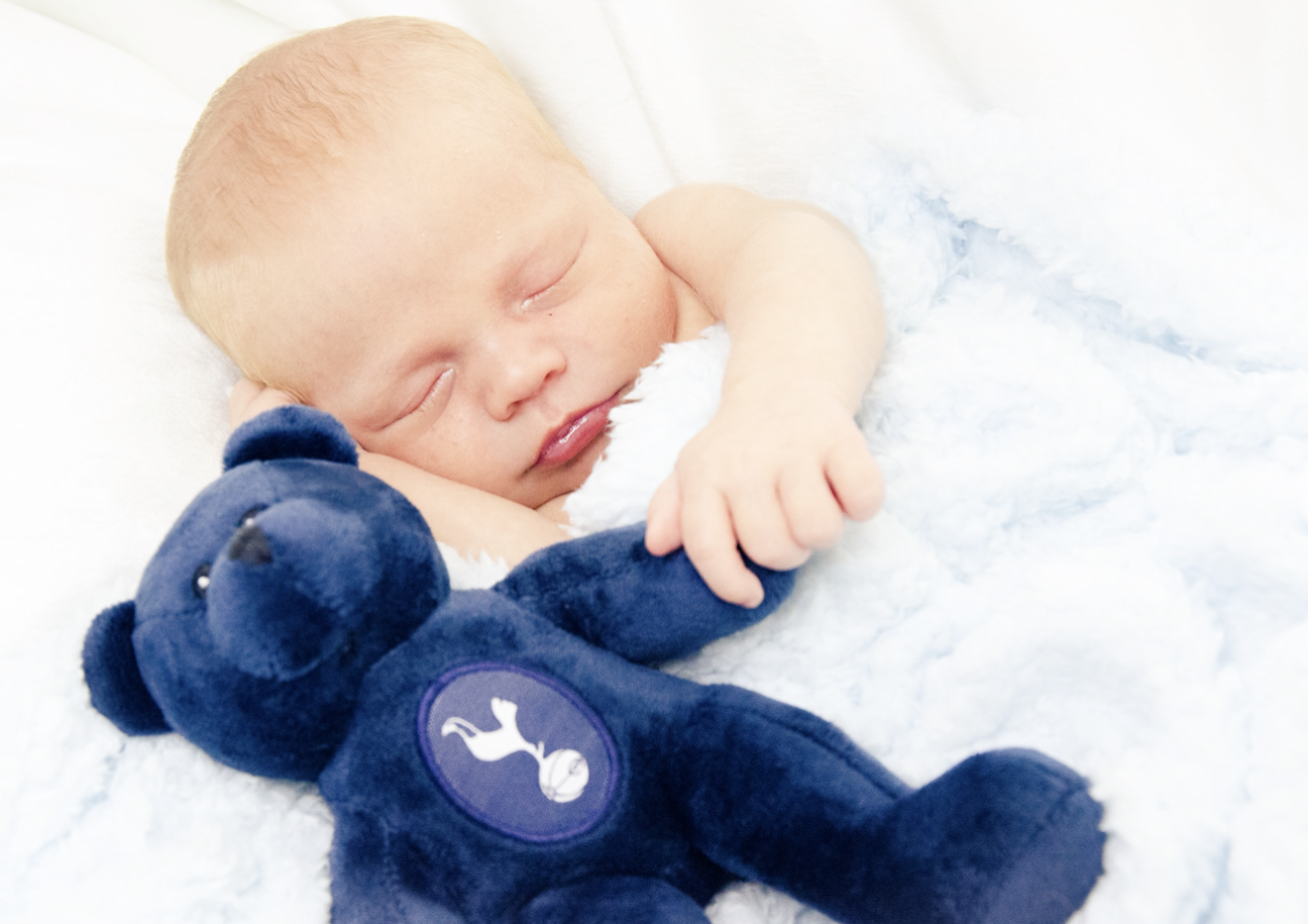 A sleeping newborn baby with light skin and red hair, resting on a white blanket, with a blue teddy bear.