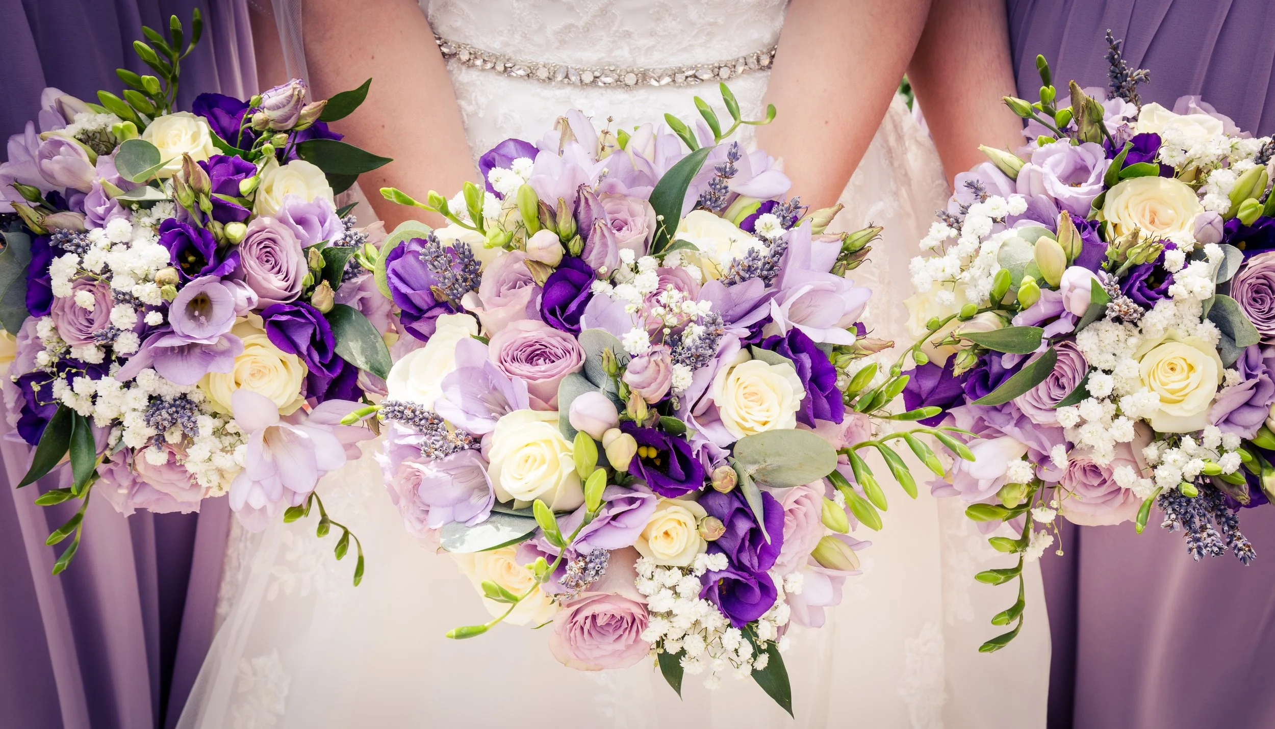 Close-up of a bride holding a large bouquet of purple, lavender, and white flowers, including roses, lisianthus, lavender, and greenery, during a wedding ceremony.