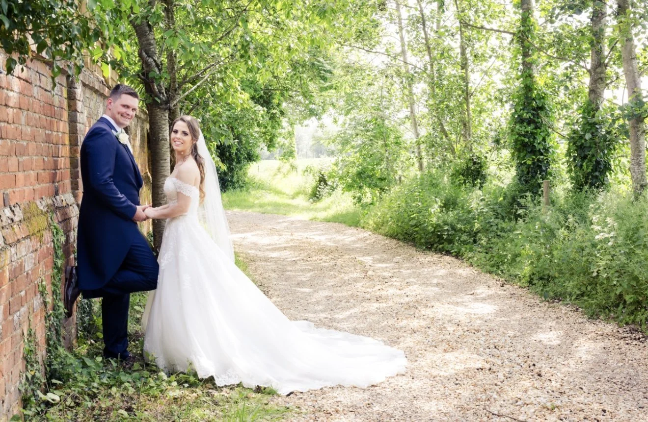 A bride and groom in wedding attire standing on a dirt path beside a brick wall and lush green trees, smiling and holding hands.