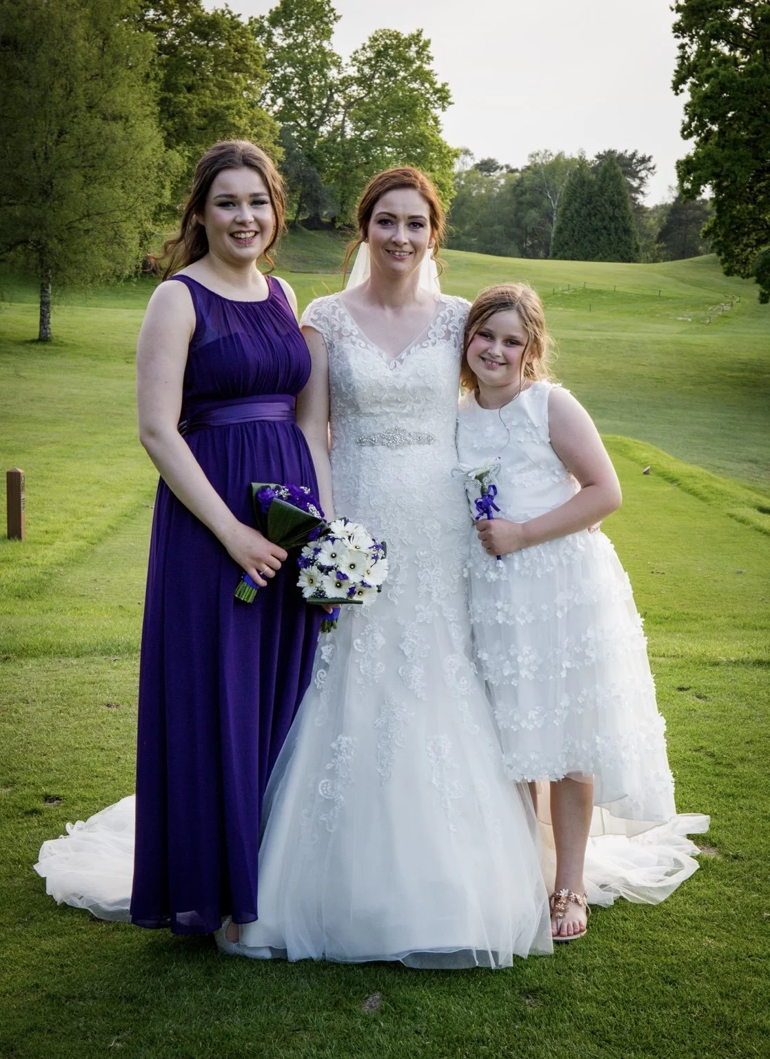 Three women standing on a grassy field in a park, one in a white wedding dress, one in a purple gown, and a young girl in a white dress, smiling and holding flowers.