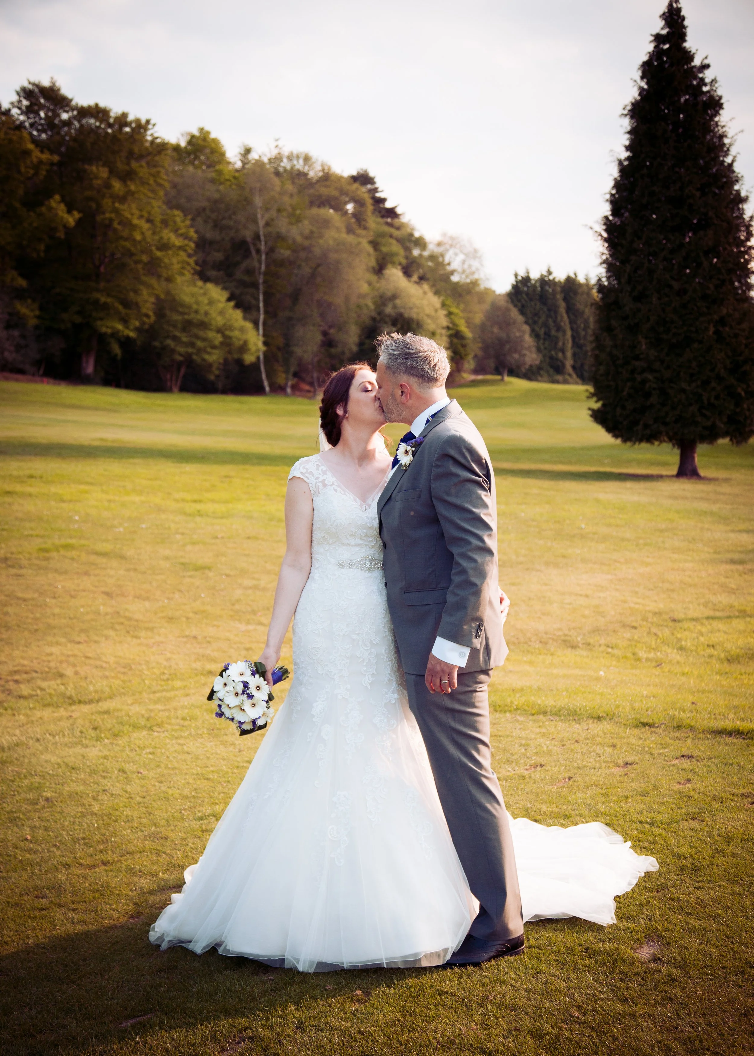 A bride and groom kissing outdoors on a grassy field with trees in the background, during sunset.