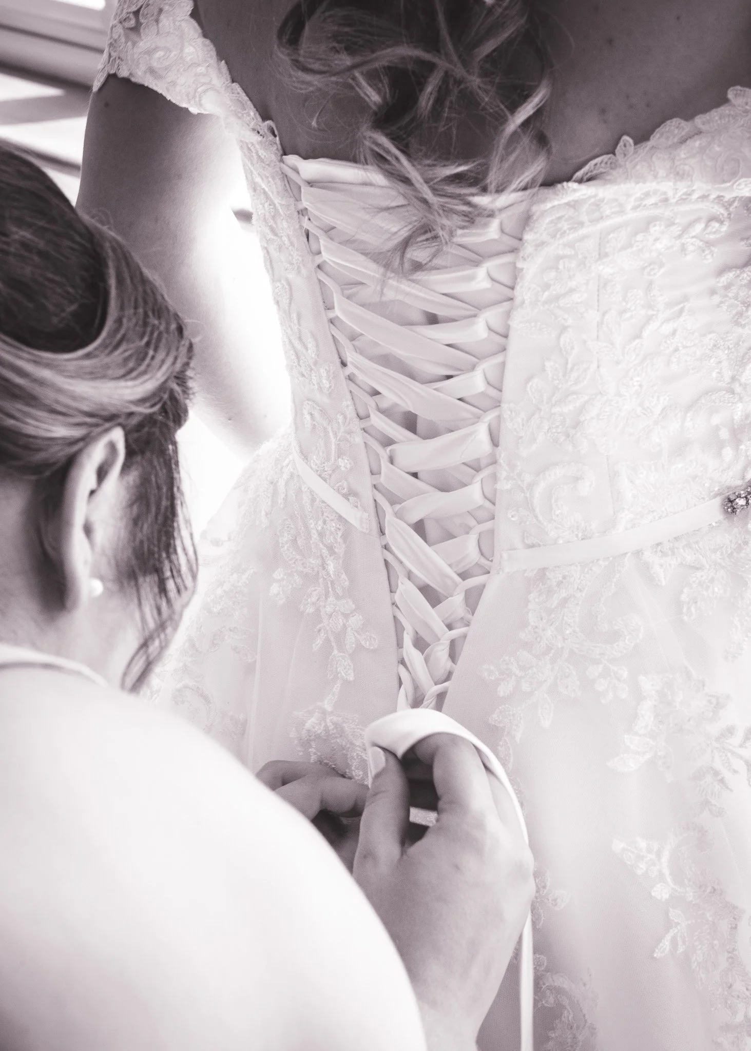 A close-up of a woman in a wedding dress laced up at the back, with a lace and embroidered fabric, as someone helps tighten the laces from behind.