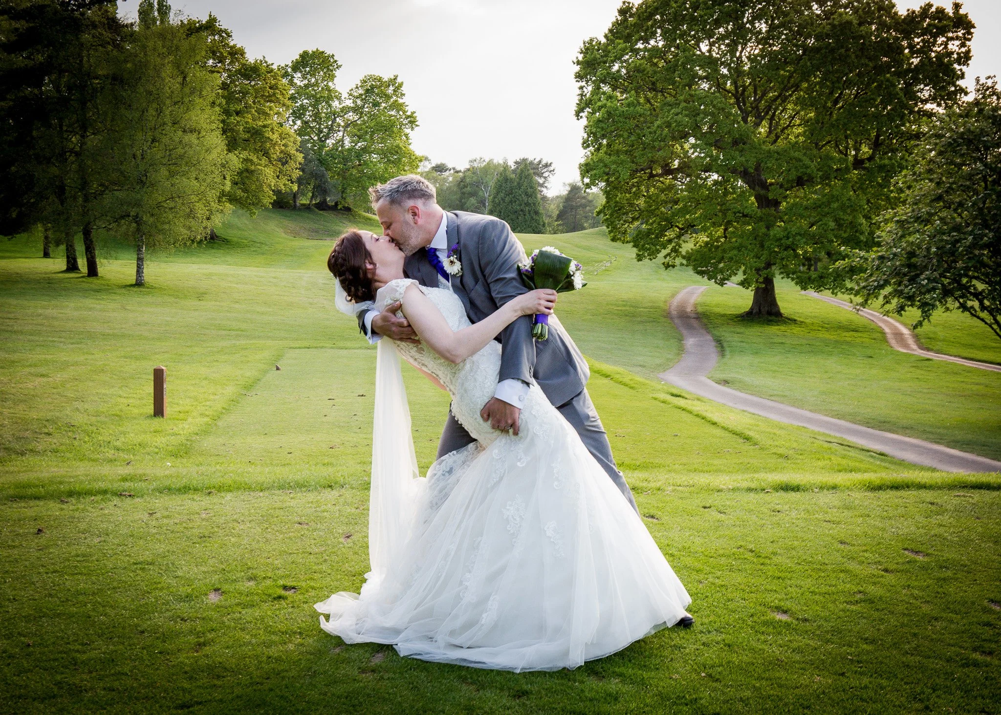 A newlywed couple kissing in a grassy park with large green trees and winding paths.