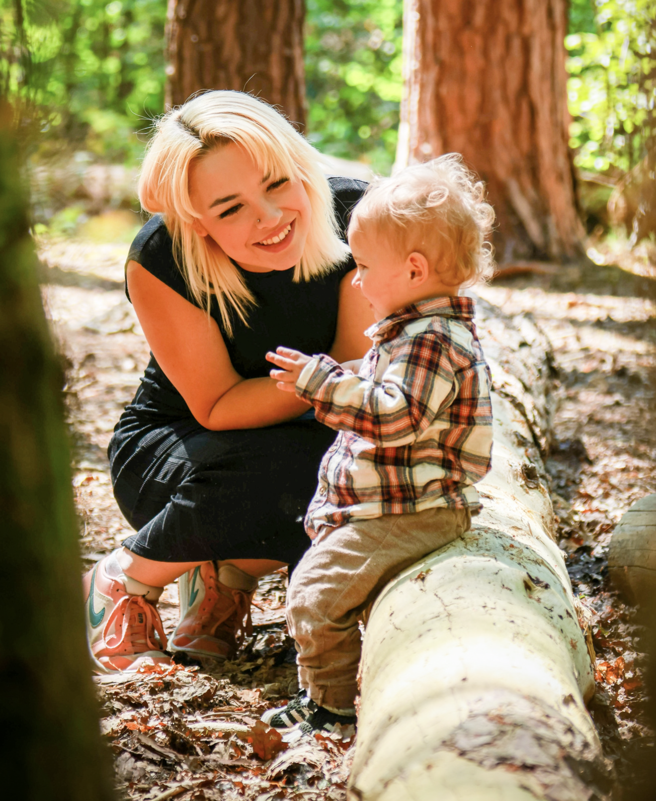 A young woman with blonde hair smiling and holding hands with a toddler boy with curly hair, sitting on a fallen tree in a forest with tall trees and green foliage.