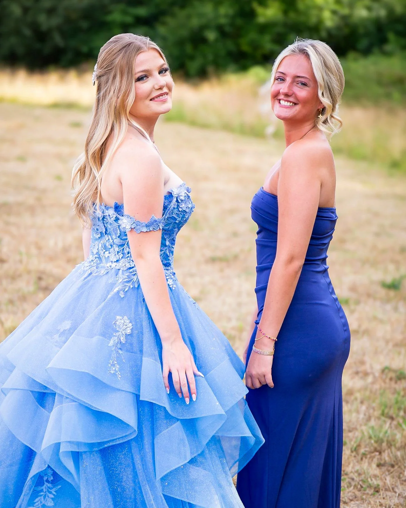 Two women standing outdoors, one in a blue ball gown and the other in a matching blue strapless dress, smiling at the camera with a natural backdrop of trees and grass.