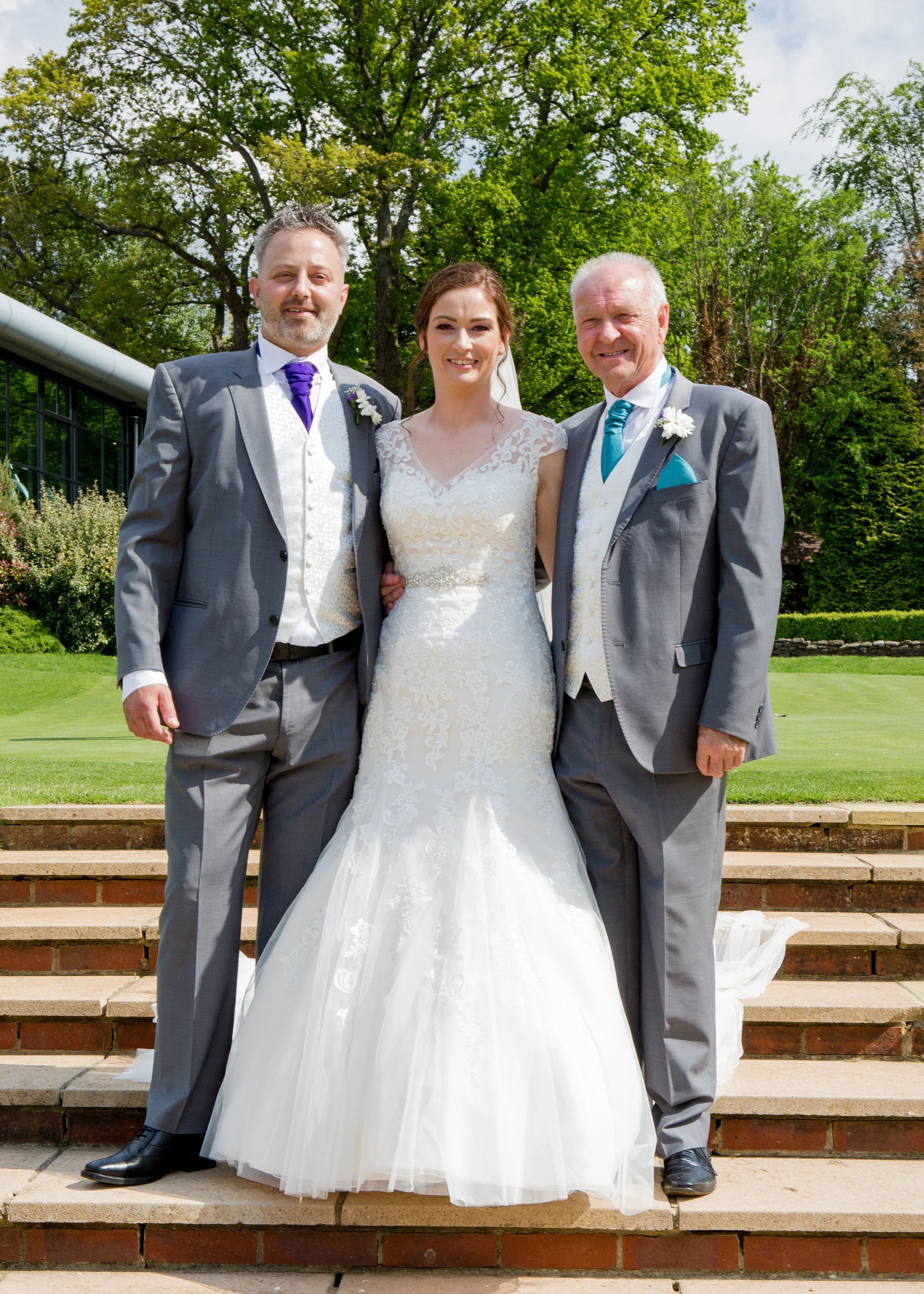 A bride in a white wedding dress standing between two men in gray suits outdoors on steps with green trees in the background.