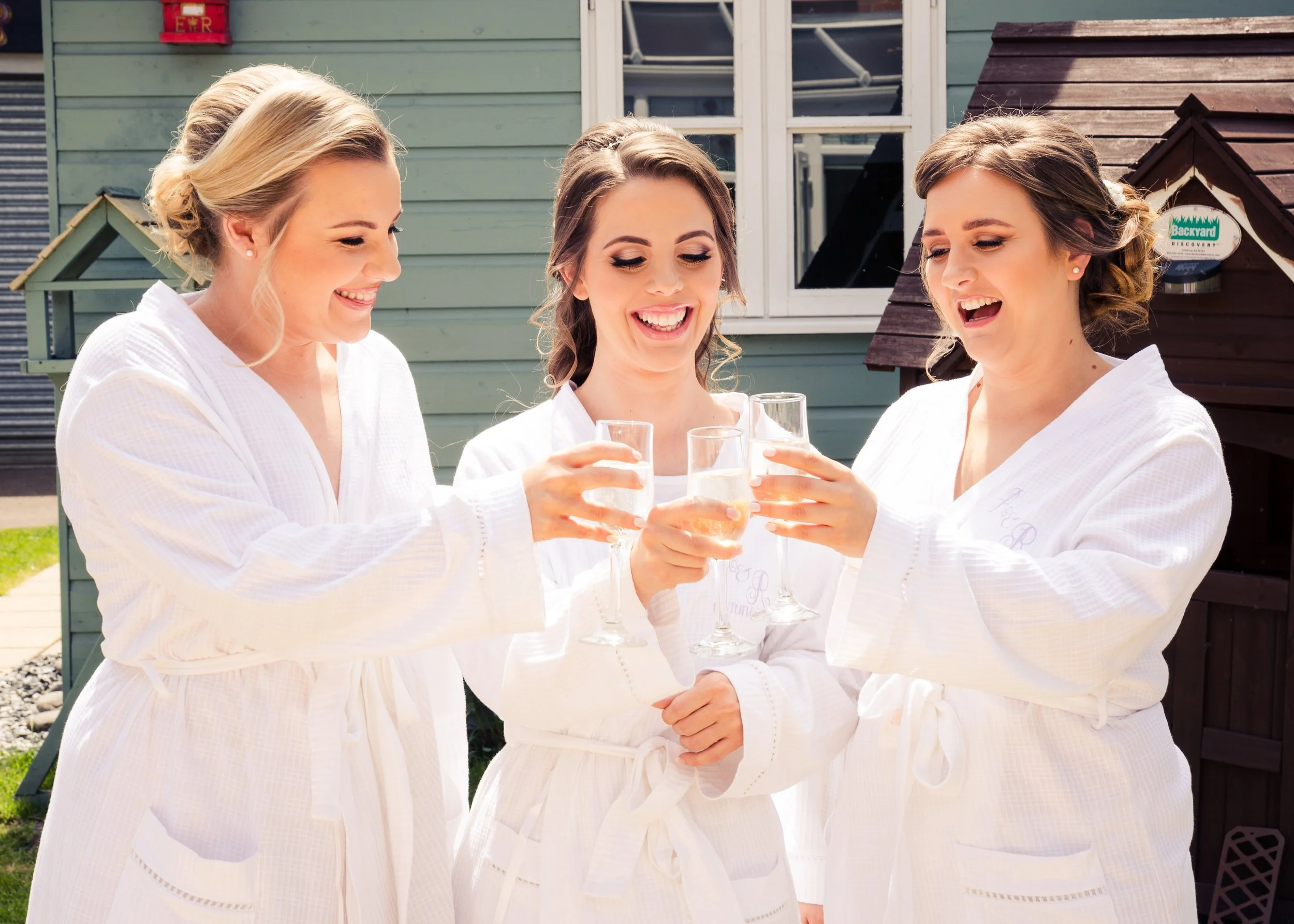 Three women in white robes celebrating with champagne outdoors in front of a house with green and dark brown siding.