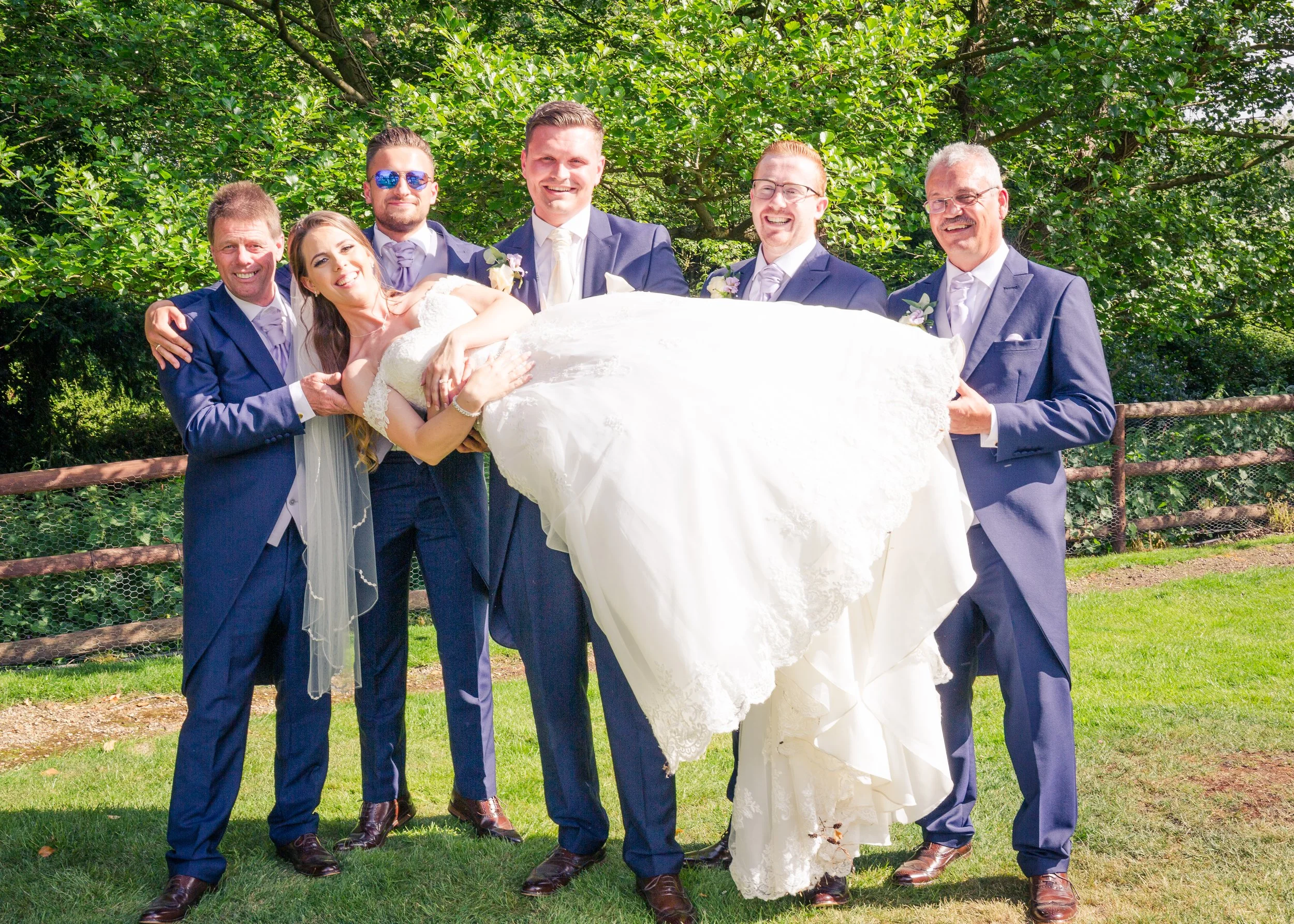 A group of seven people, including six men in suits and one woman in a wedding dress, outdoors on a sunny day with a green leafy background. They are smiling and posing, holding the bride who is lying across the arms of the men.