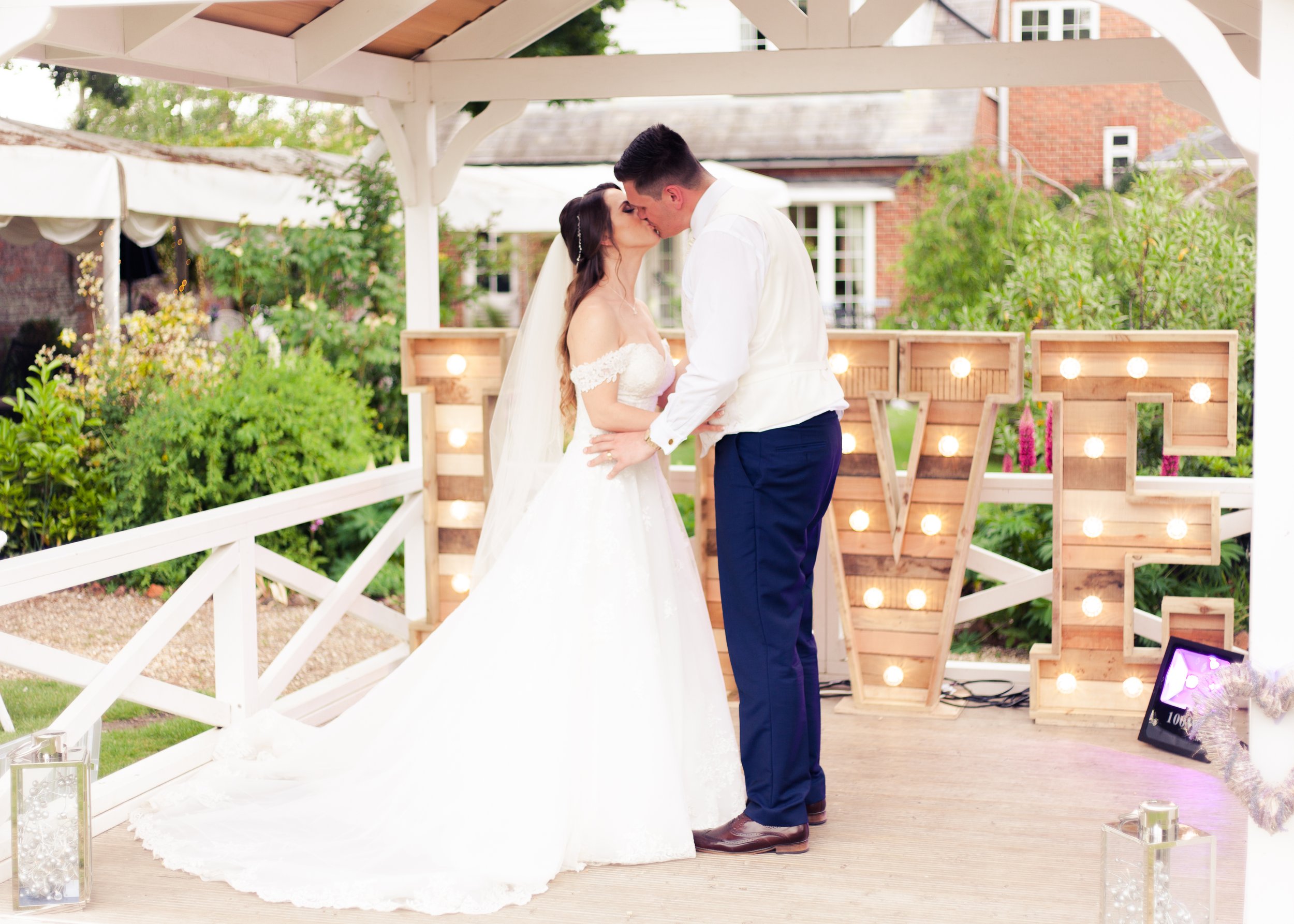 A bride and groom sharing a kiss during their wedding ceremony outdoors, under a white wooden gazebo decorated with Edison bulb lights and surrounded by green plants.