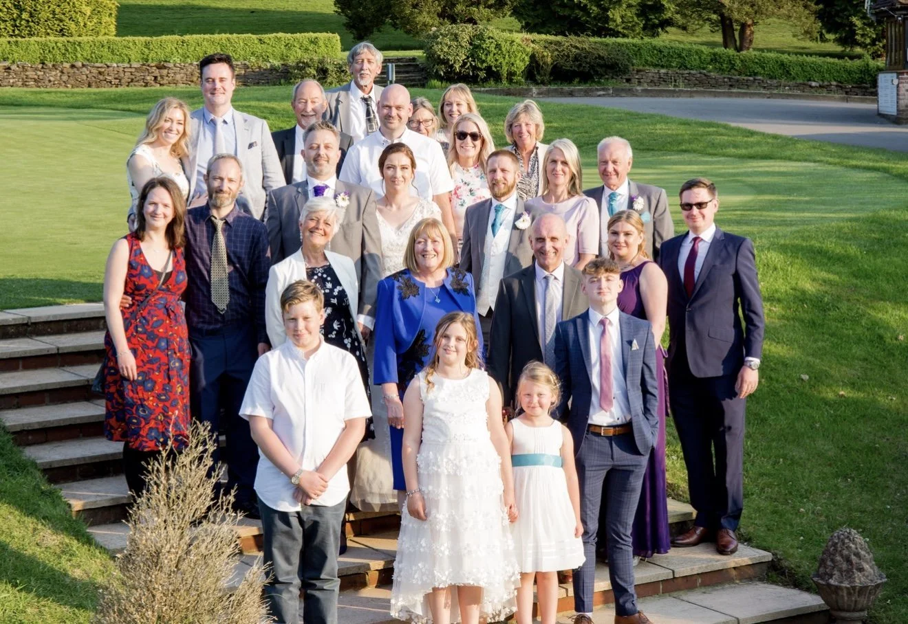 A large group of people dressed in formal attire posing outdoors on steps with a grassy background, likely at a wedding or celebration.