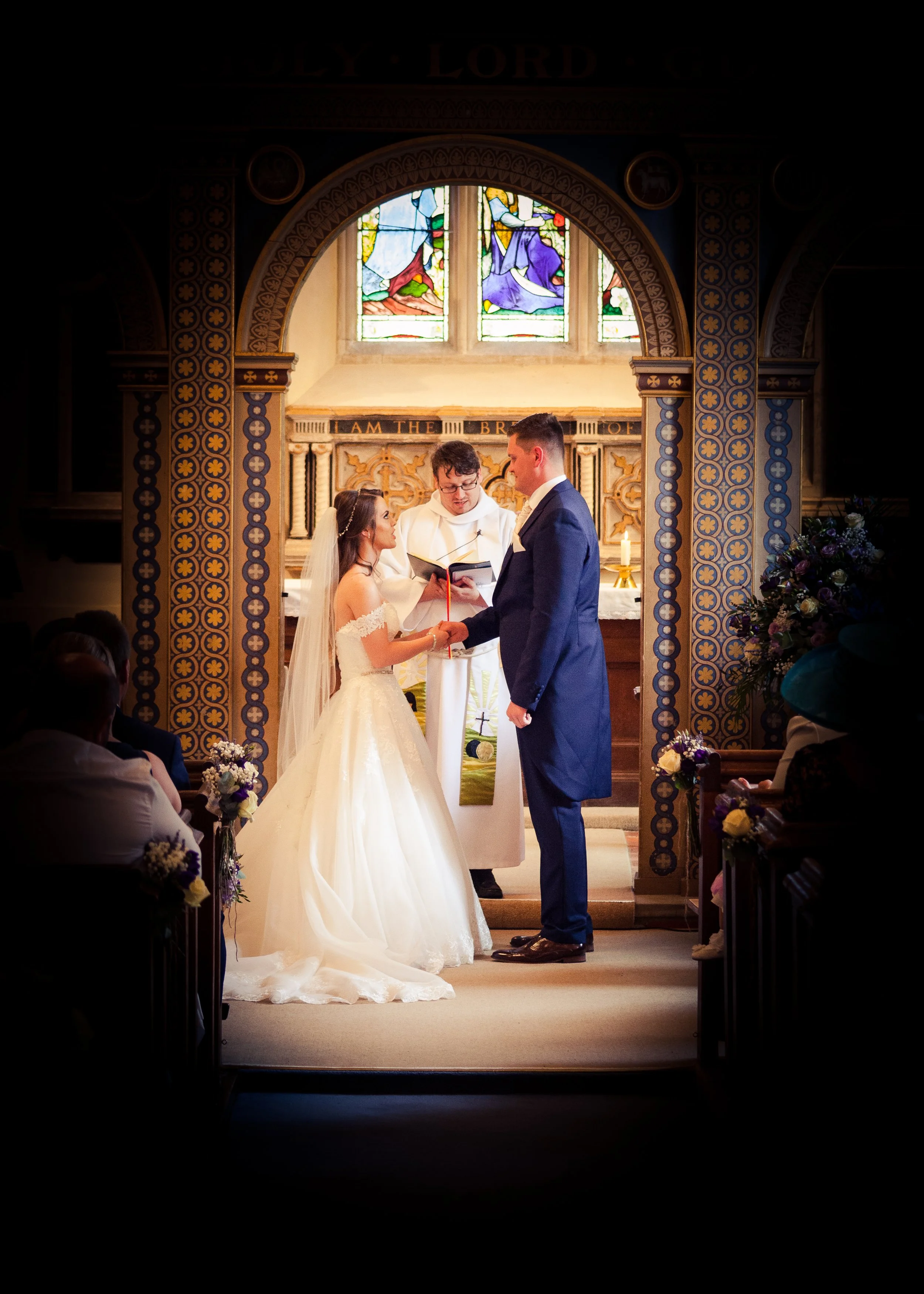 A couple getting married in a church, holding hands and looking at each other, with a priest officiating. There are stained glass windows behind them and guests seated on either side.