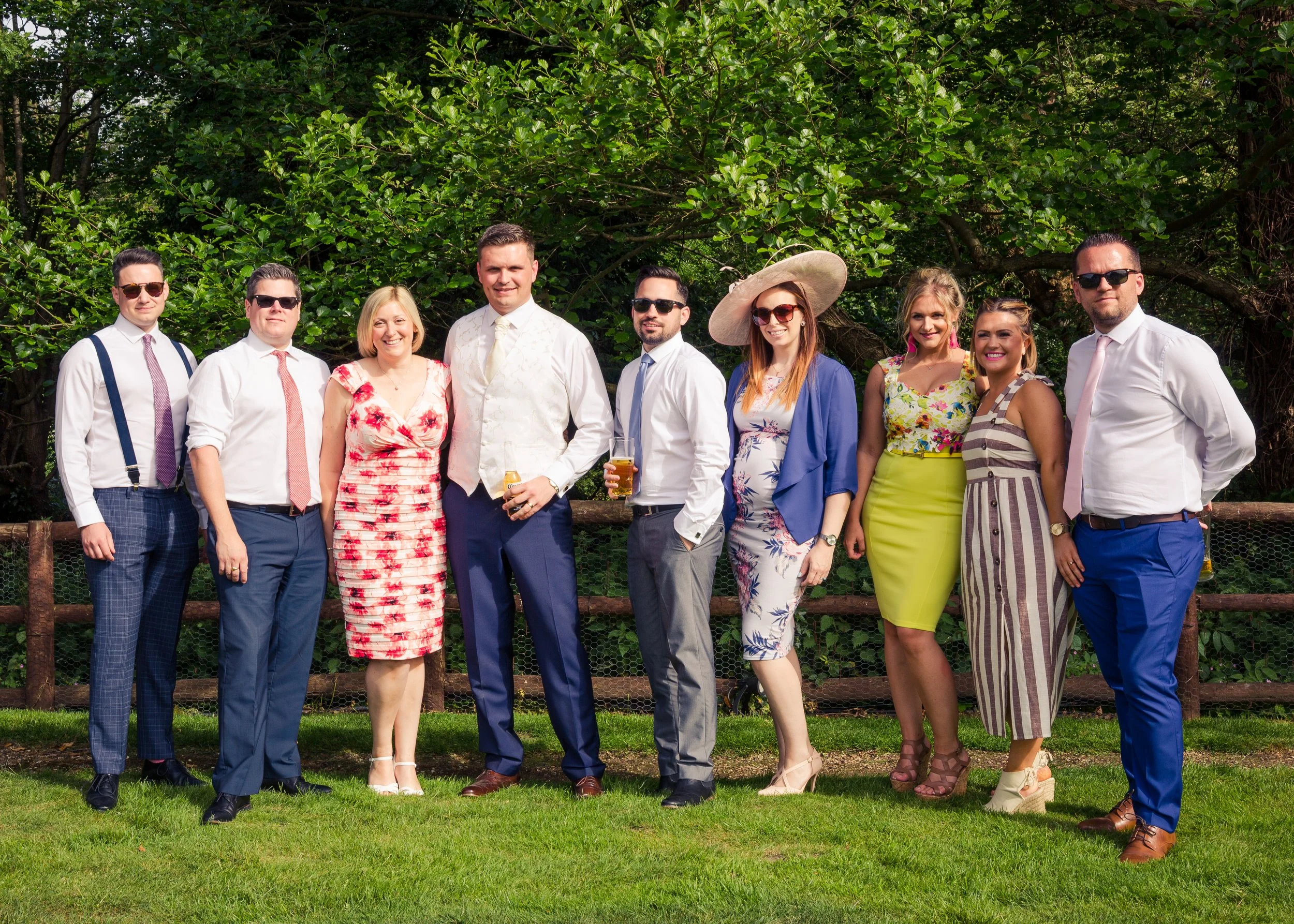 A group of nine people dressed in colorful and formal clothing, standing outdoors on a grassy area with trees in the background, smiling at the camera, some holding drinks.