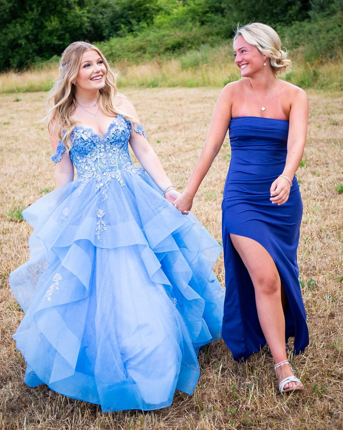 Two women holding hands and smiling outdoors, one in a blue ball gown, the other in a strapless blue gown with a slit, standing in a grassy field with trees in the background.