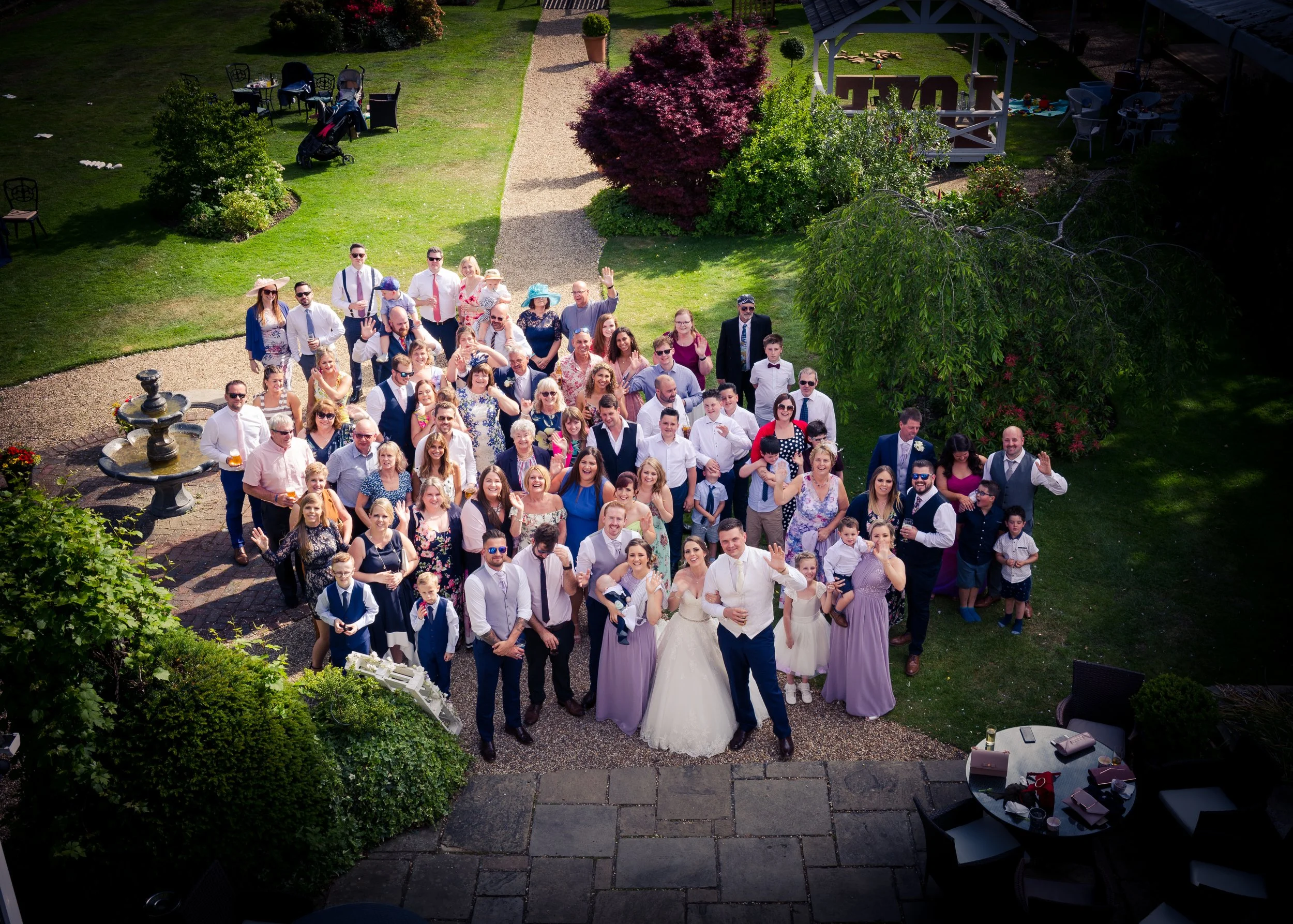 A large group of people posing for a photo outdoors at a celebration, including men, women, and children, some dressed in formal attire including a bride in a wedding dress and groom in a suit. The setting is a lush garden with trees, bushes, and a g