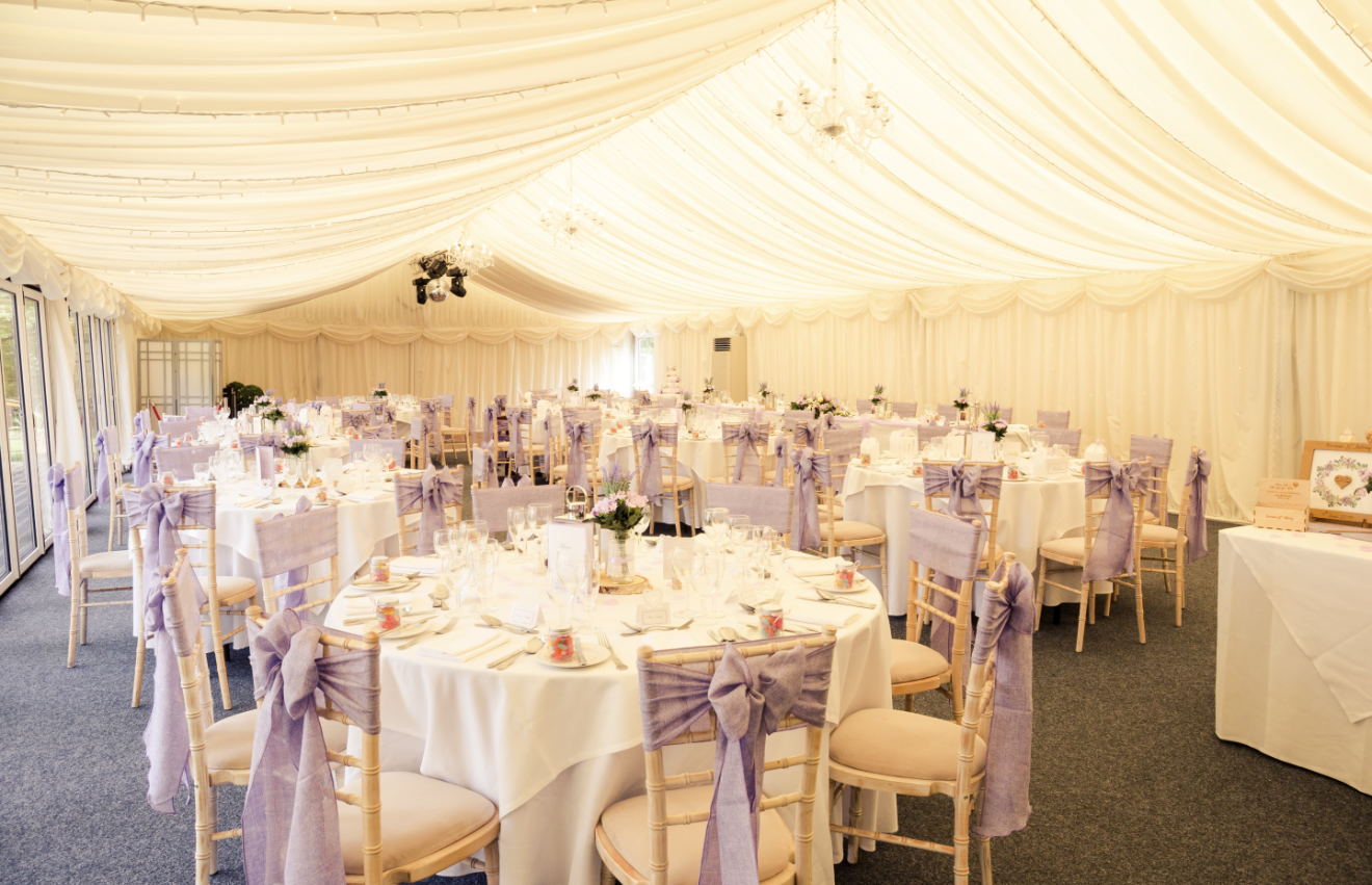 Decorated event tent with round tables covered in white tablecloths, lavender chair sashes, floral centerpieces, and place settings, illuminated by chandeliers and sunlight.