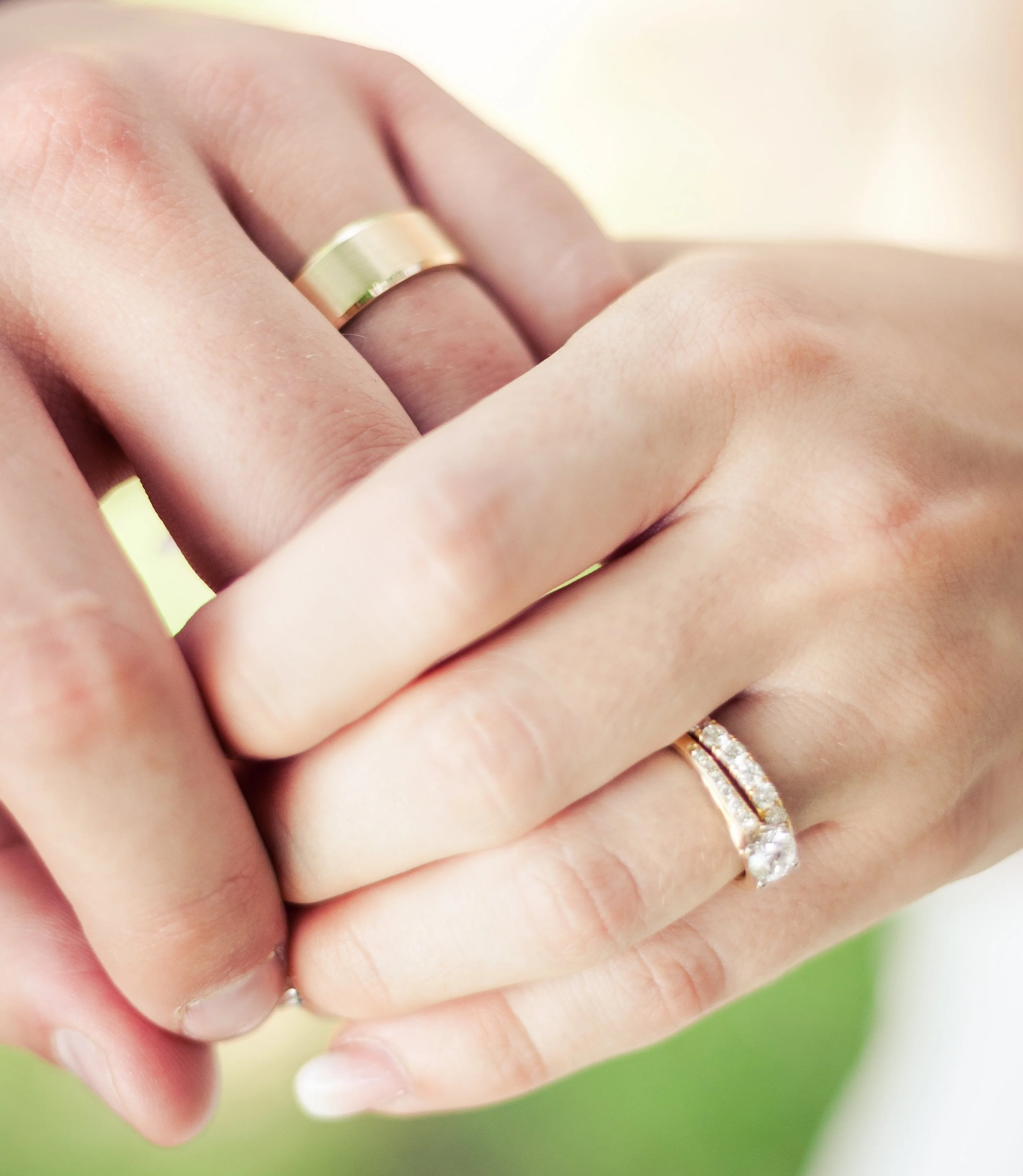 Close-up of a person's hand wearing a gold wedding band and an engagement ring with diamonds, against a blurred background.
