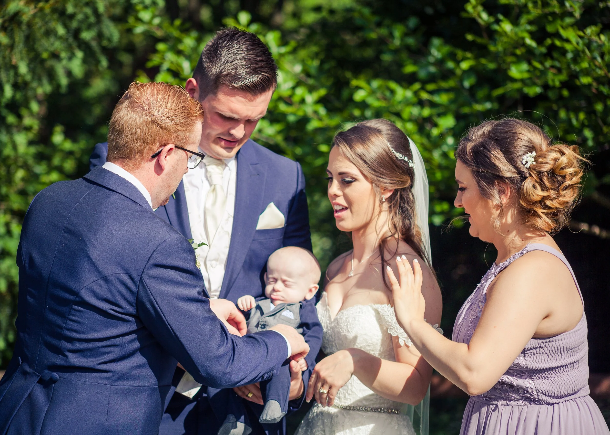 A wedding ceremony outdoors with a couple exchanging vows, surrounded by family members including children, against a backdrop of green trees.