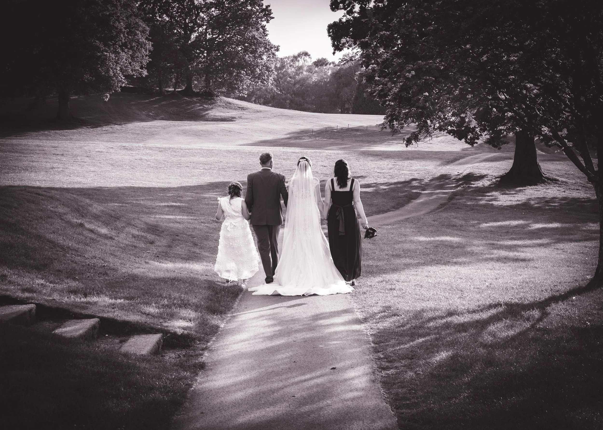 A group of four people, including a bride wearing a wedding dress and veil, a groom in a suit, a woman in a black dress, and a young girl in a white dress, walking together on a golf course surrounded by trees.