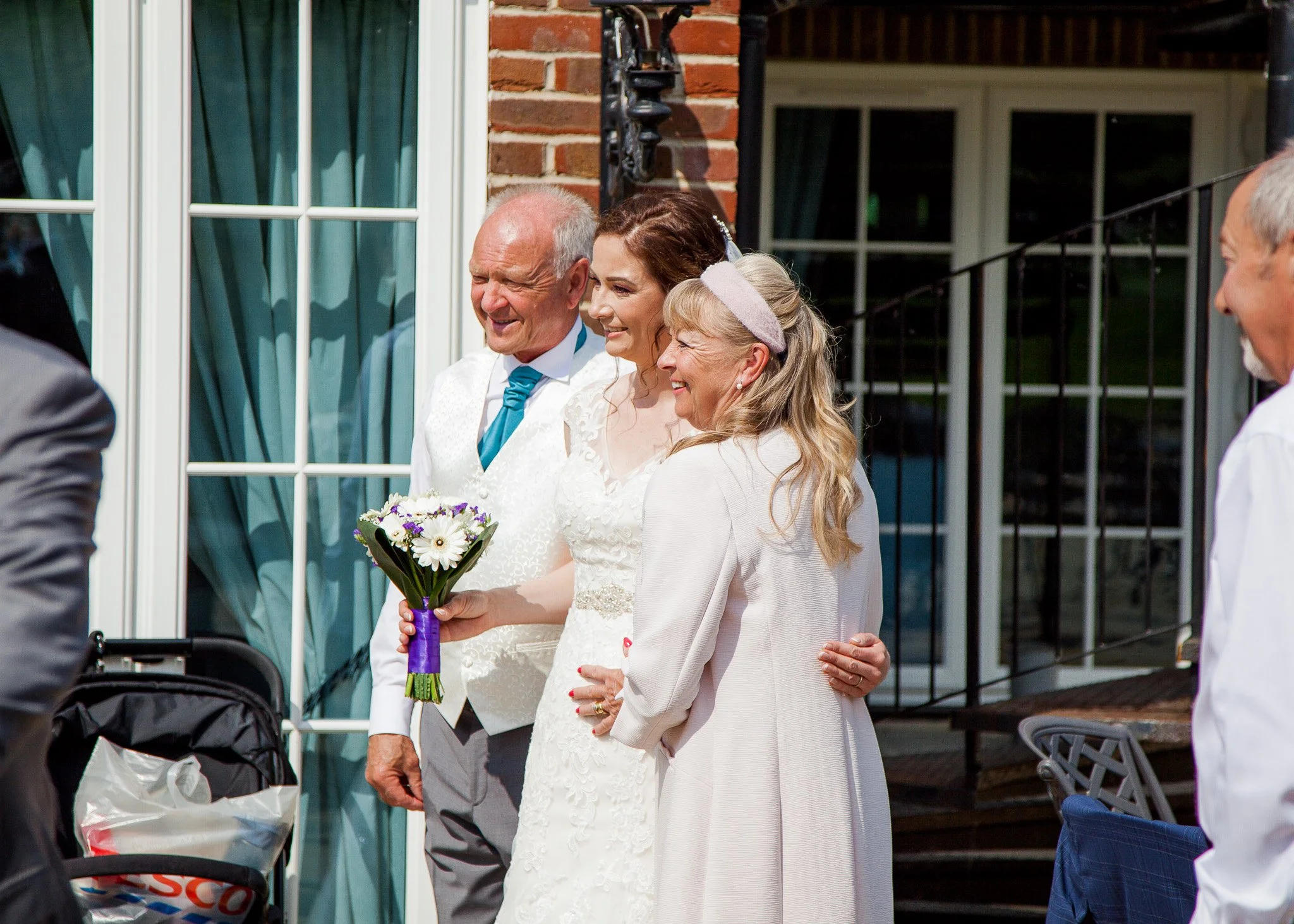 A bride holding a bouquet of flowers standing next to an older woman, possibly her mother, at an outdoor wedding. An older man with gray hair and a wedding suit is also visible, smiling nearby.