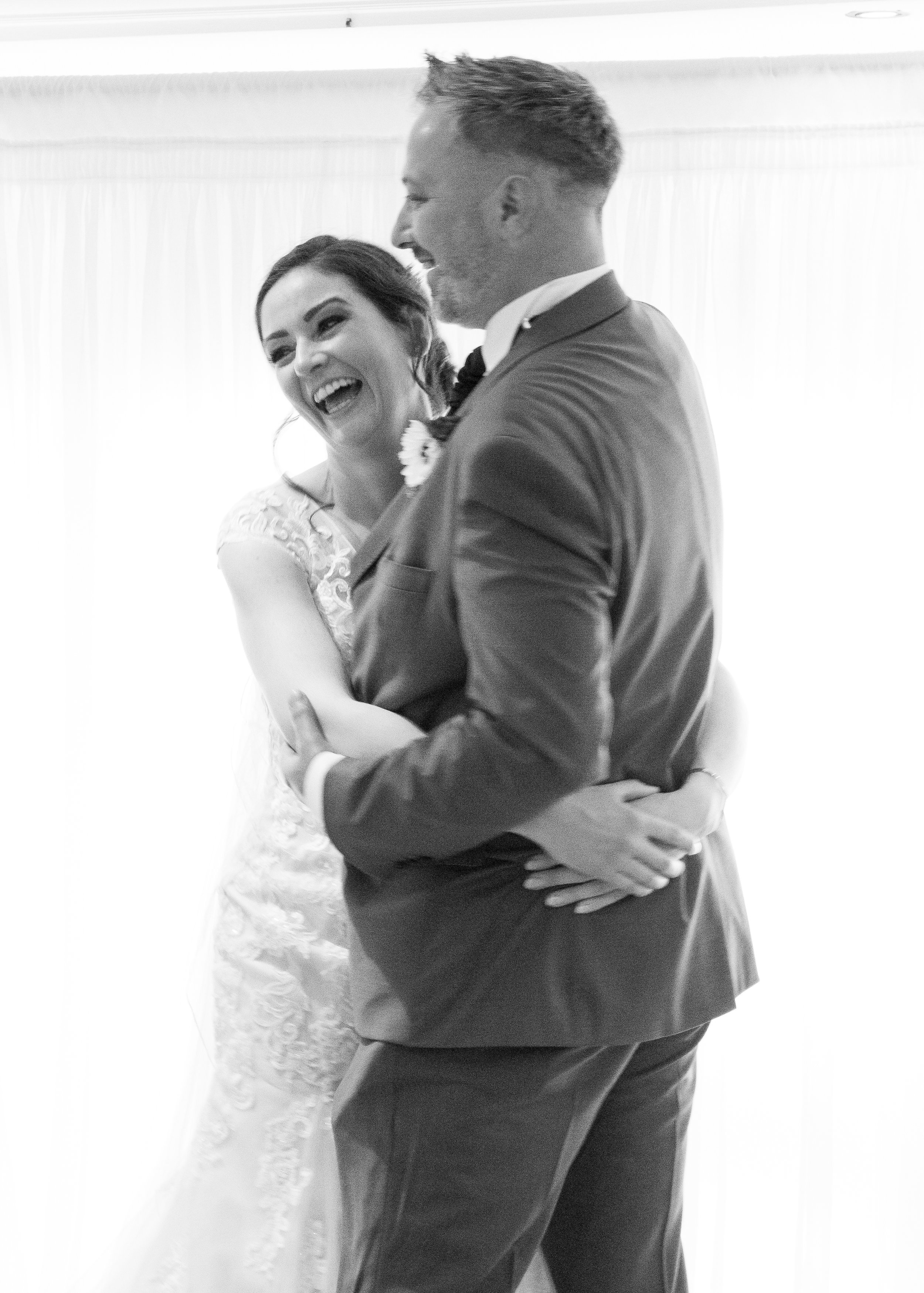 A couple dancing closely together, smiling and sharing a joyful moment, dressed in formal wedding attire, in a bright room with curtains.