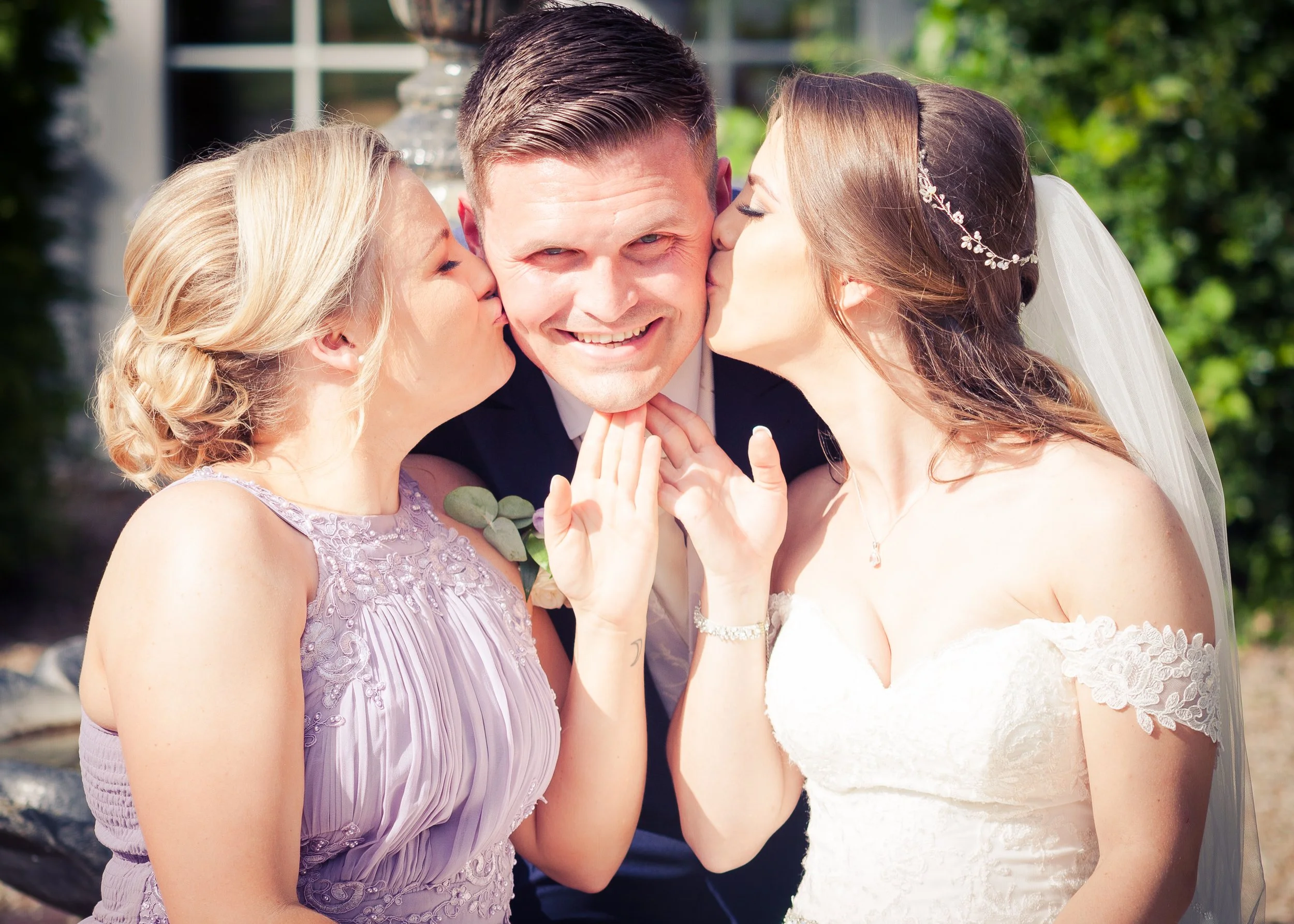 Two women kissing a man on each cheek at a wedding, with the man smiling in the center. The woman on the right is wearing a wedding dress and veil, the woman on the left is wearing a lavender dress, and the man is in a dark suit. Greenery and a windo