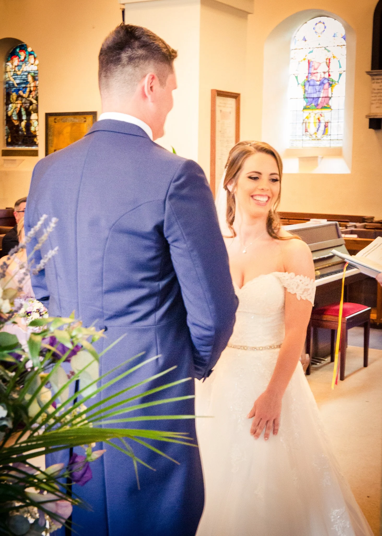 A bride and groom standing in a church during a wedding ceremony. The groom is wearing a blue suit, and the bride is wearing a white wedding gown. The bride is smiling while looking at the groom.