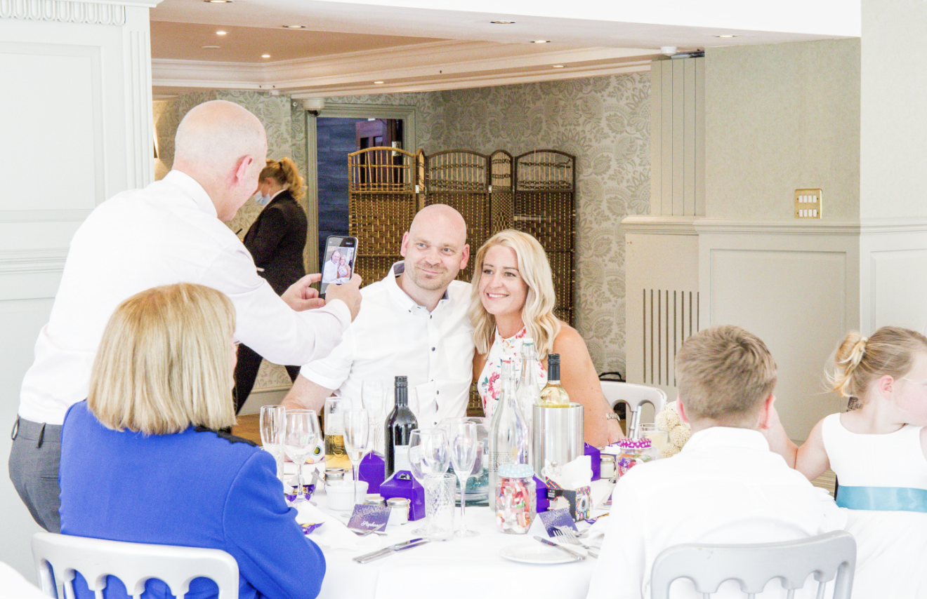 People taking a photo of a couple at a dinner table in a well-decorated room.