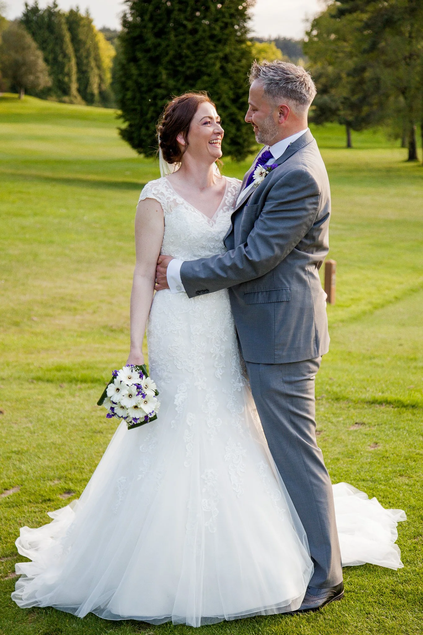 A bride and groom stand close in an outdoor setting with green grass and trees, smiling and gazing into each other's eyes. The bride is wearing a white lace wedding gown and holding a bouquet of white and purple flowers. The groom is dressed in a gra