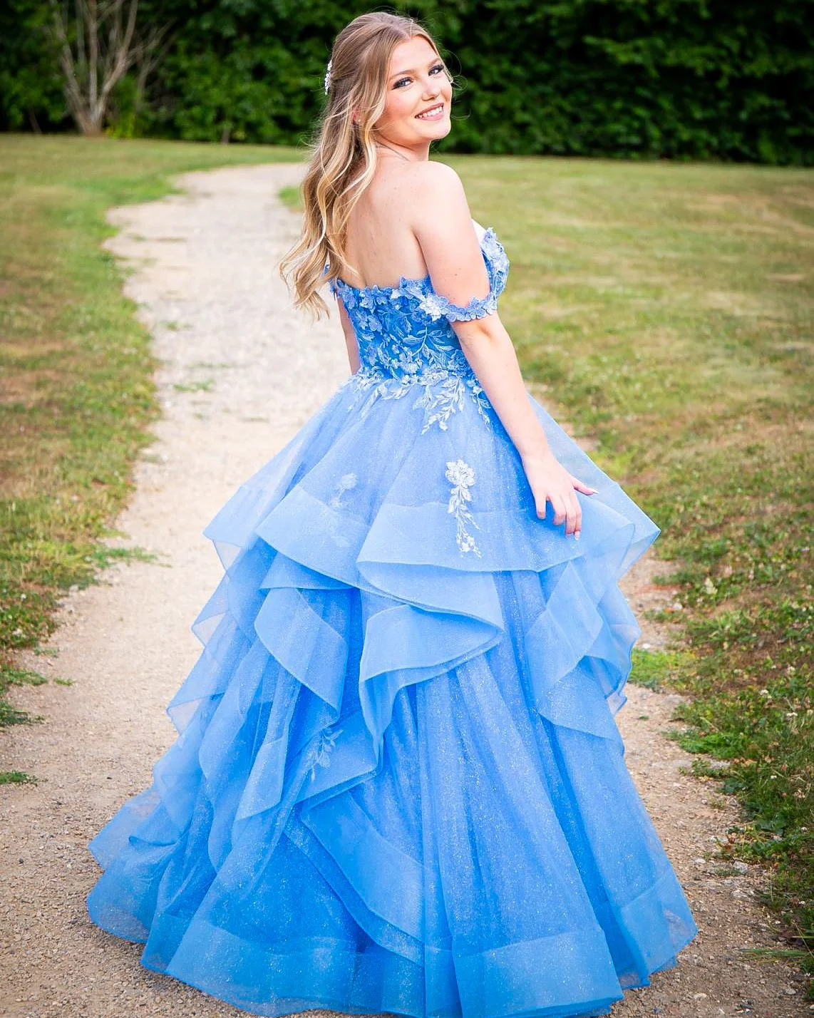 Young woman in a blue, off-the-shoulder, layered gown standing outdoors on a dirt path with green grass and trees in the background, smiling at the camera.