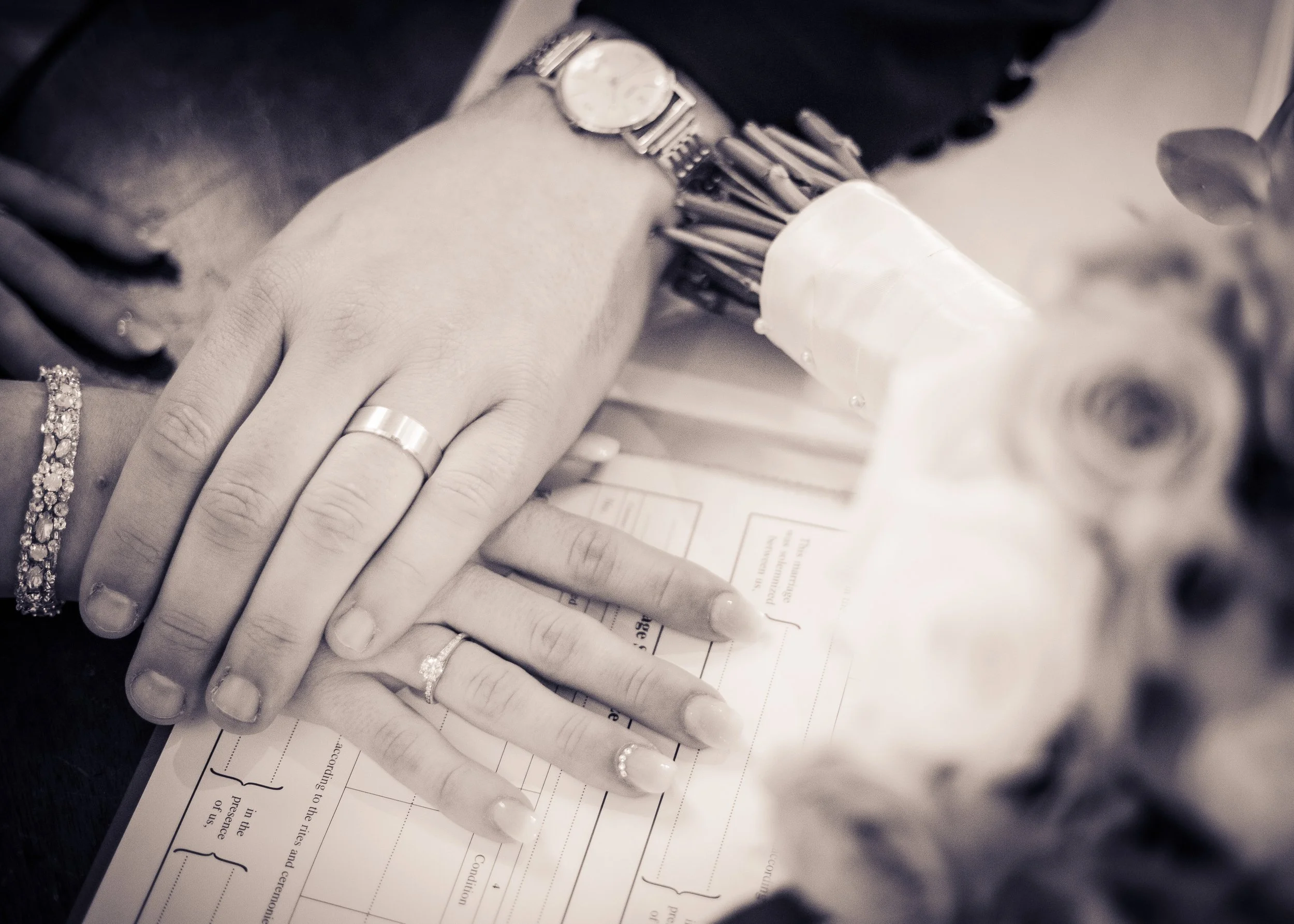 Close-up of a person's hand wearing rings and a watch, resting on a document with printed text, a pen, and other objects nearby.