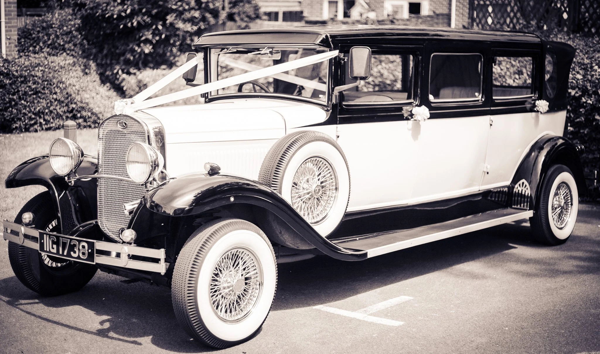 A vintage black and white car decorated with ribbons and flowers, parked on a street with greenery in the background.