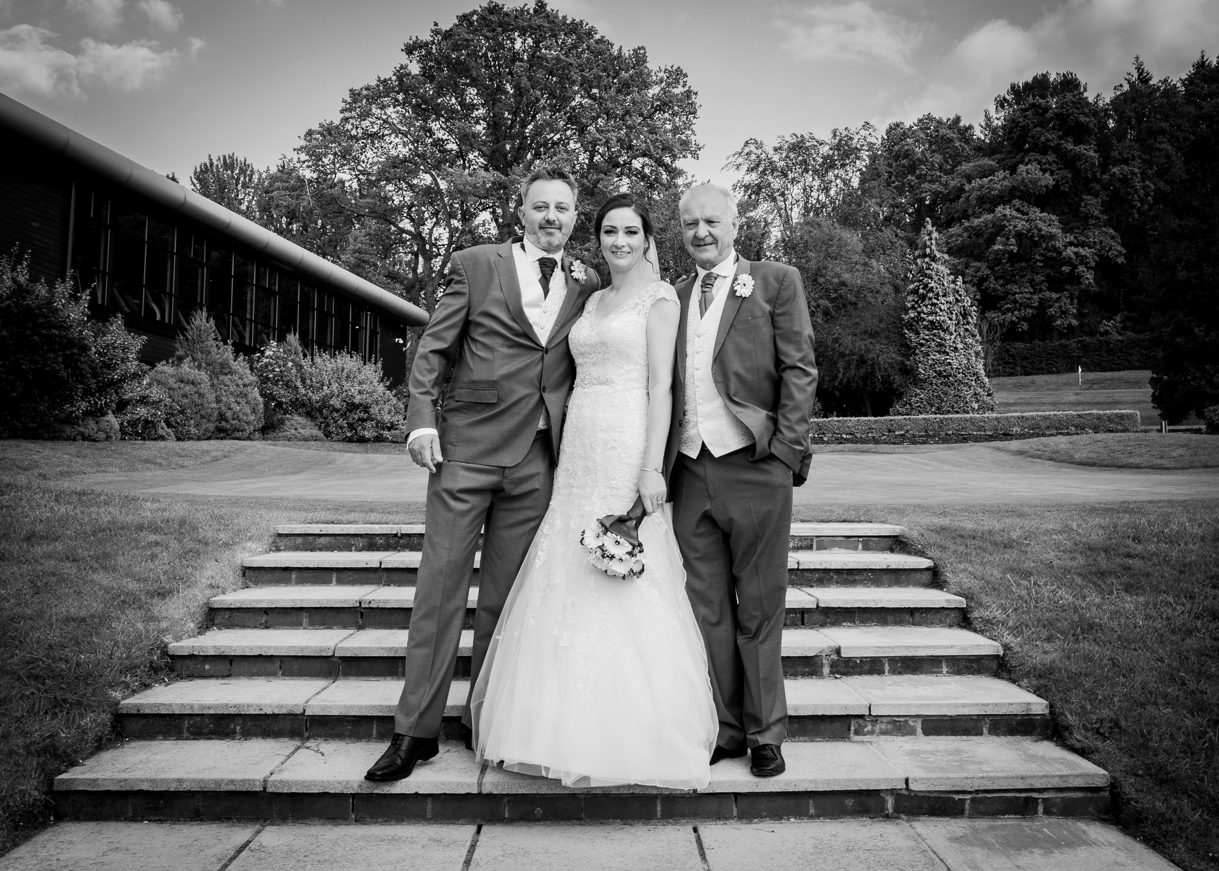 Black and white photograph of a bride and two men standing on steps outdoors, with a building and trees in the background. The bride wears a white wedding dress, holding a bouquet, and smiling. The men wear suits with boutonnieres, and are standing c