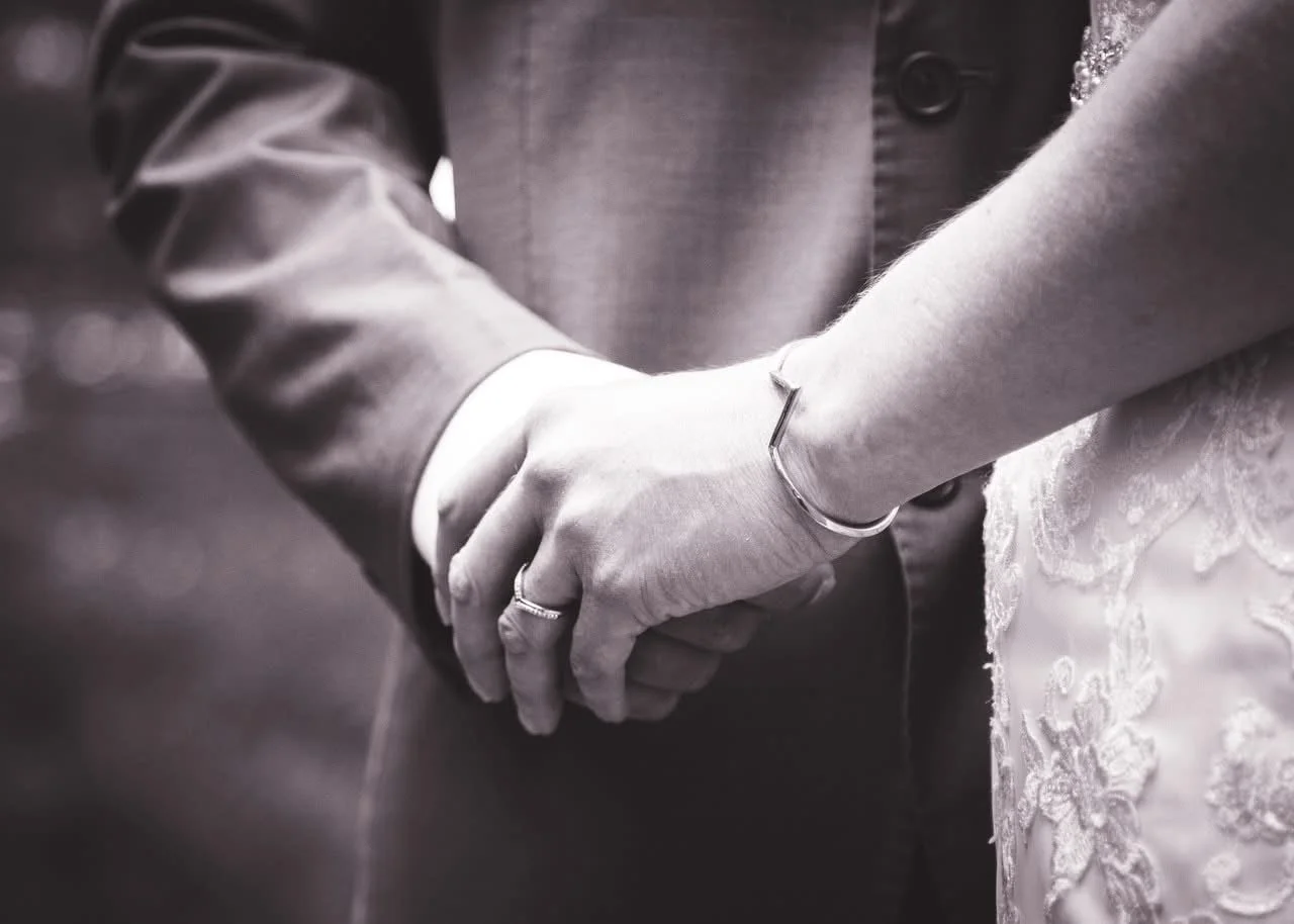 Close-up of a couple holding hands, with wedding rings visible, during a wedding ceremony in black and white.