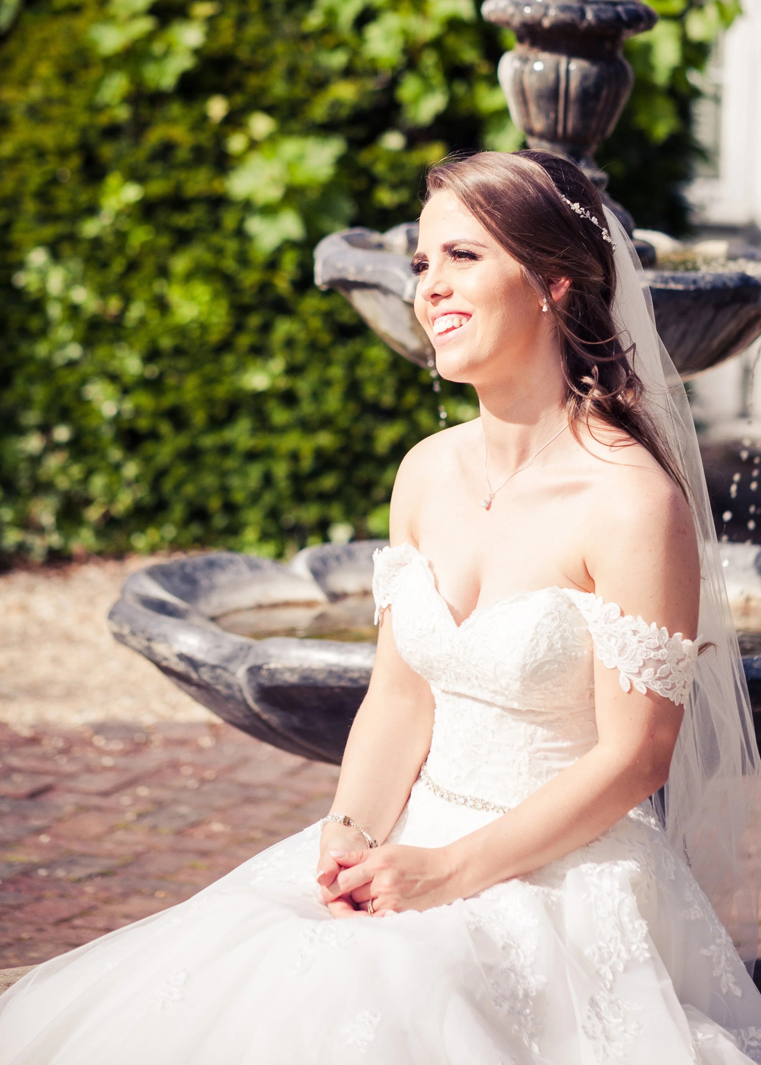 A smiling woman in a wedding dress sitting outdoors near a fountain with greenery in the background.