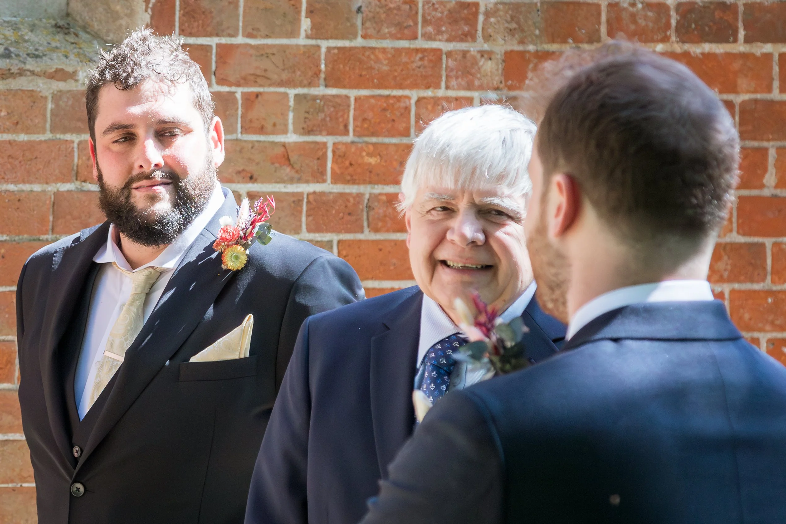 Three men in suits at an outdoor event against a brick wall, one man with white hair is smiling, and the others have brown hair, one with a beard and another facing away from the camera.