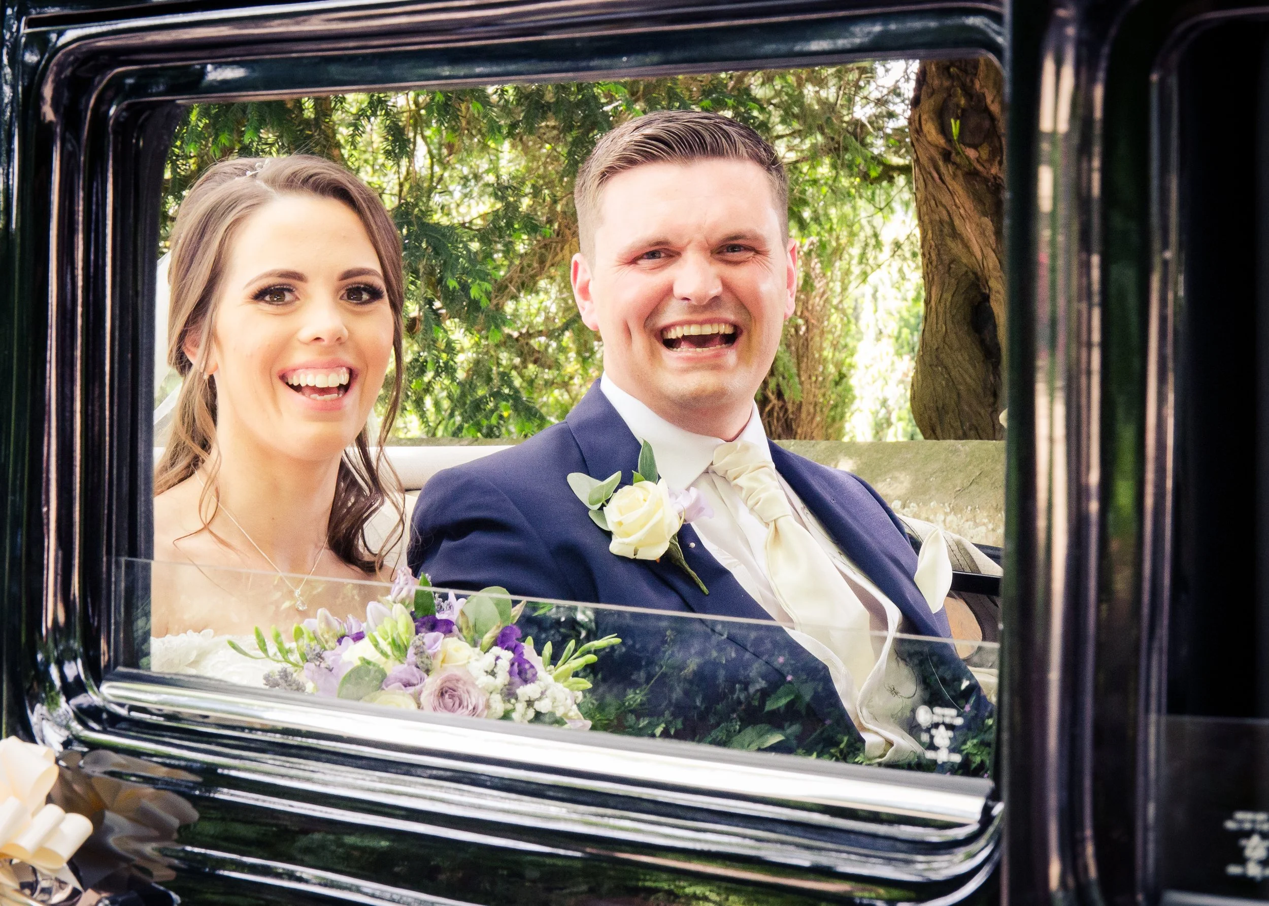 A newlywed couple smiling and sitting in a vintage car, surrounded by lush green trees, with the bride holding a bouquet of flowers.