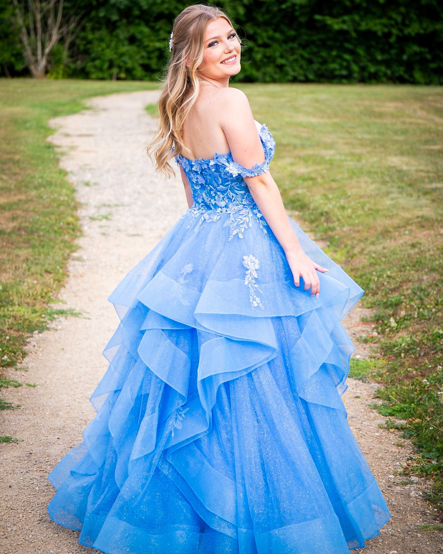 A young woman in a blue, off-shoulder princess gown with floral embroidery and layered tulle skirt, standing outdoors on a gravel path with green grass and trees in the background, smiling at the camera.