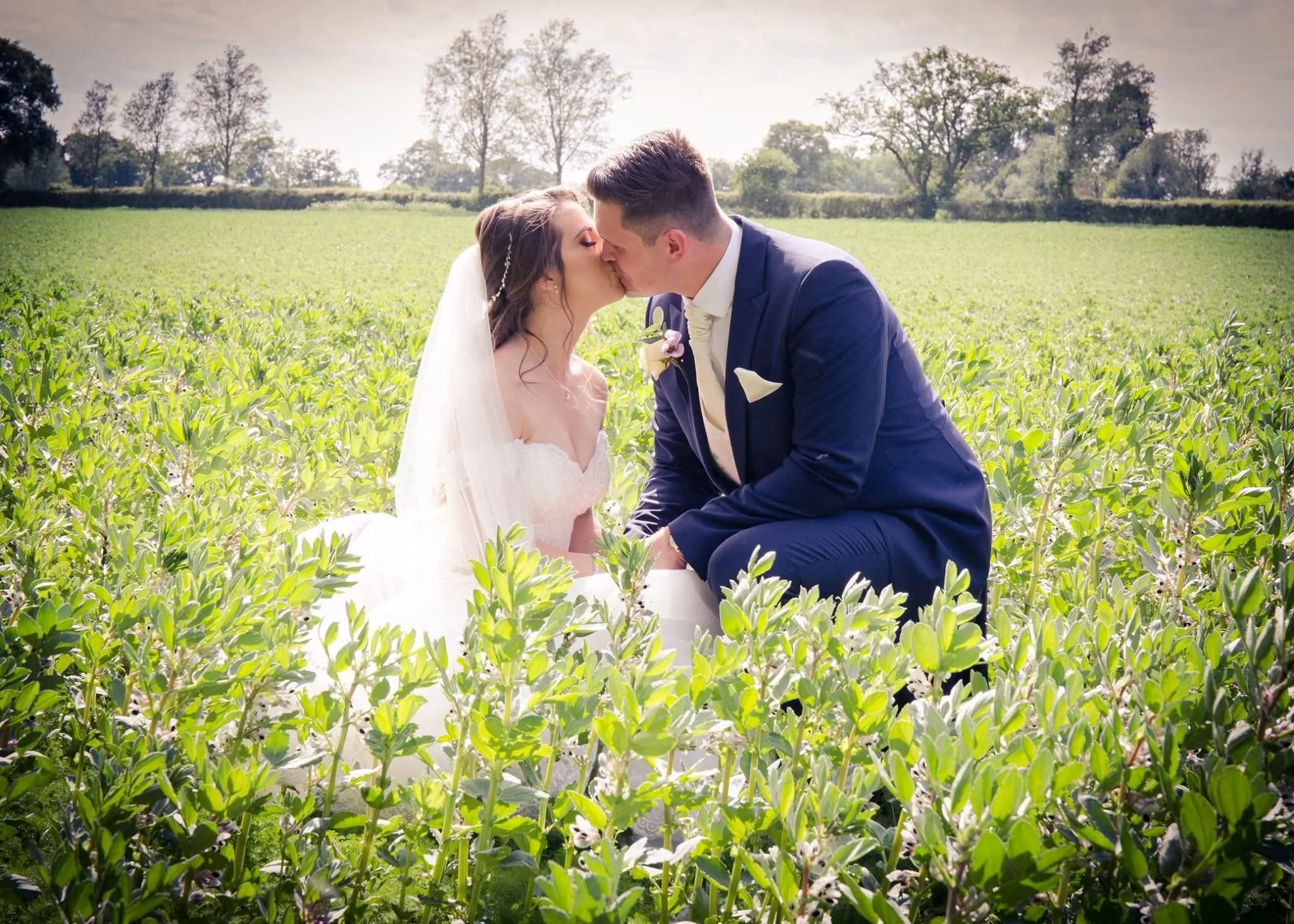 A bride and groom kissing in a field of green plants with trees and a partly cloudy sky in the background.