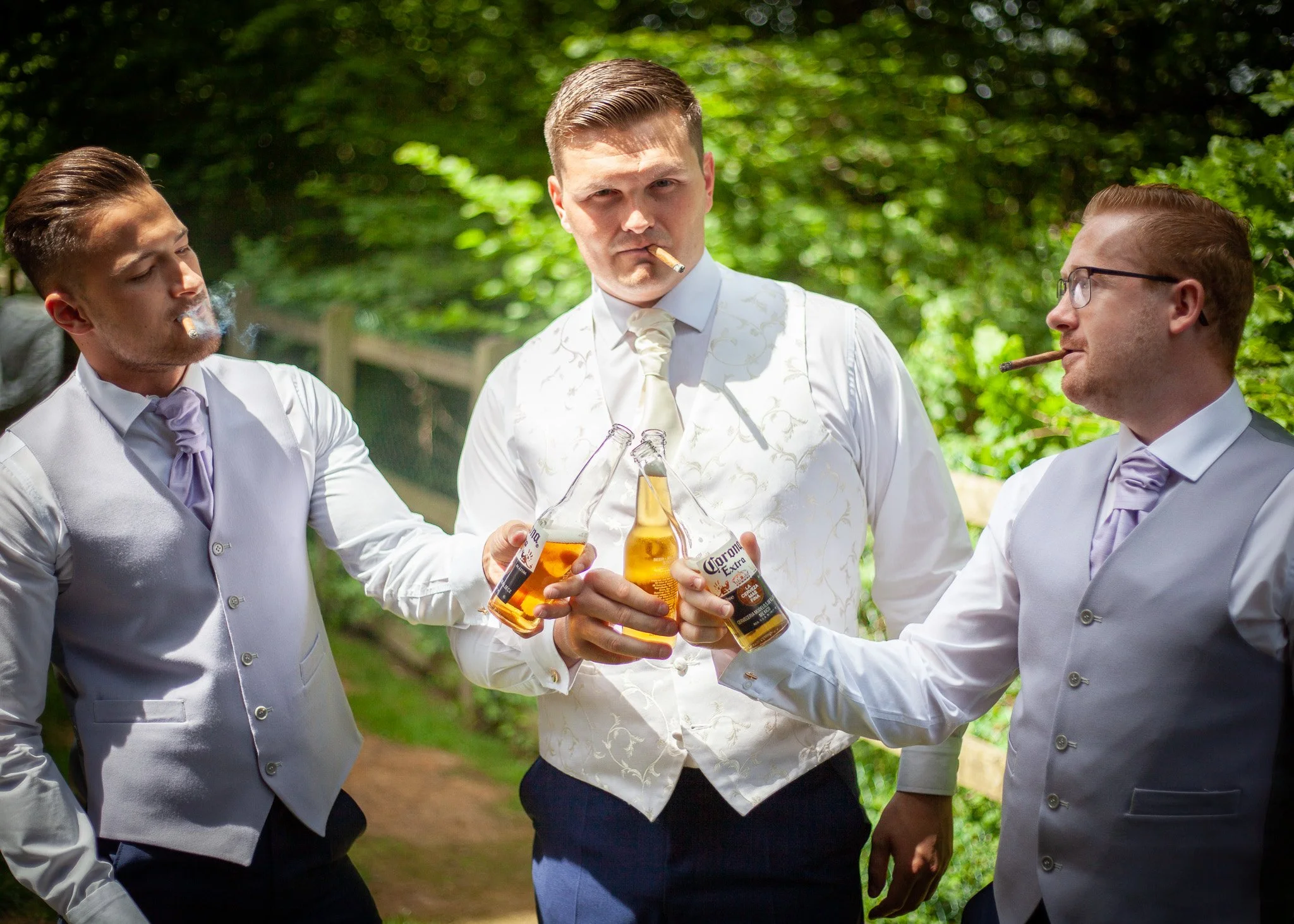 Three men in formal suits holding beer bottles, outdoors in a green natural setting, with two of them smoking cigars.