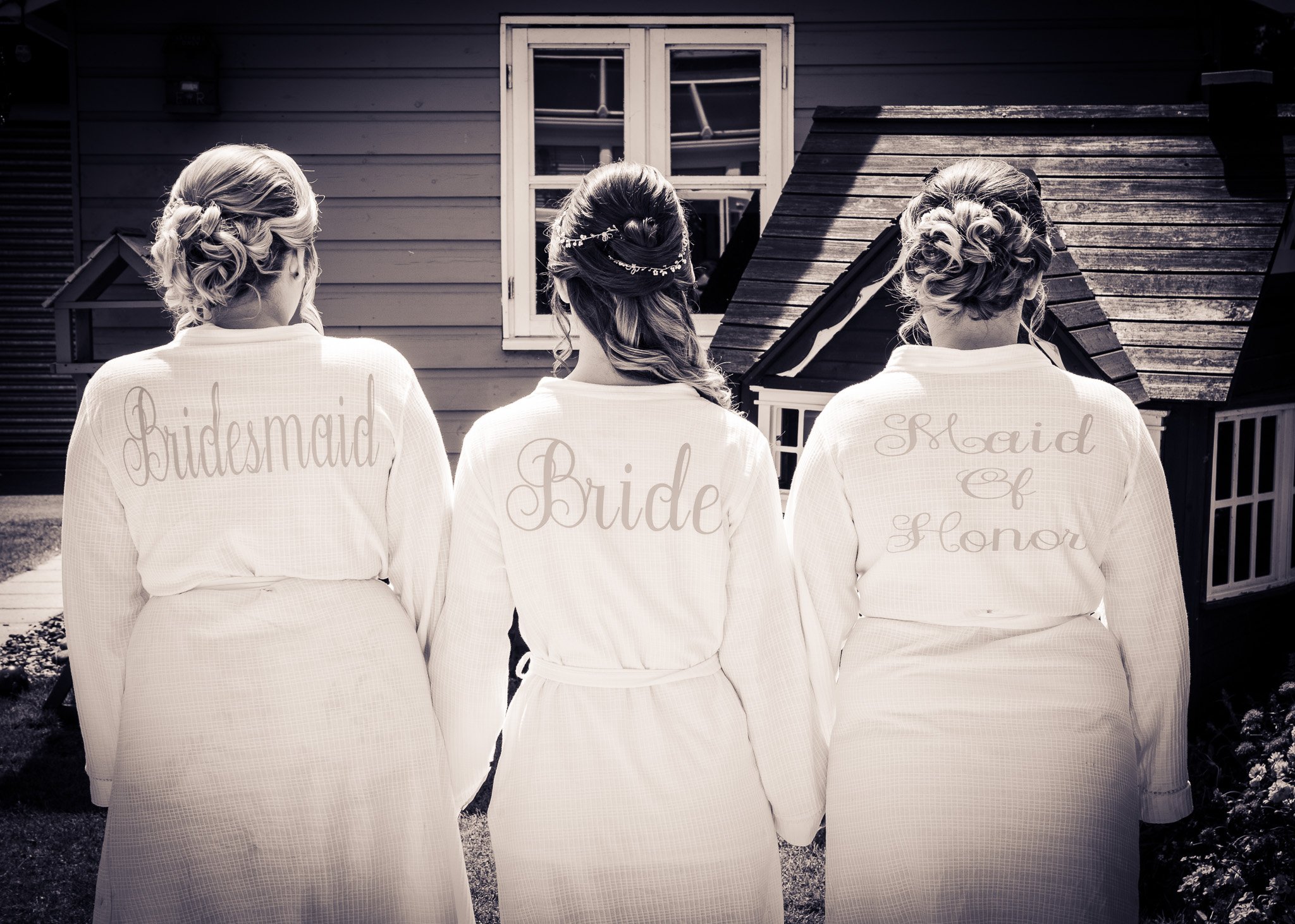 Three women dressed as bridesmaids with their backs to the camera, standing outdoors in front of a house, wearing robes that read 'Bridesmaid', 'Maid of Honor', and 'Bridesmaid'.