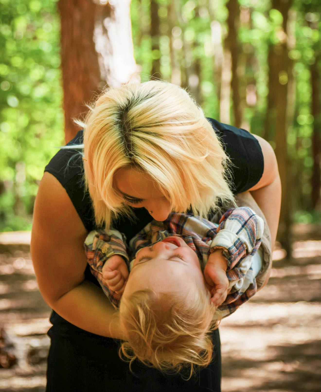 A woman with blonde hair is holding a young child in a forest, and the woman is leaning down to kiss or nuzzle the child's face.
