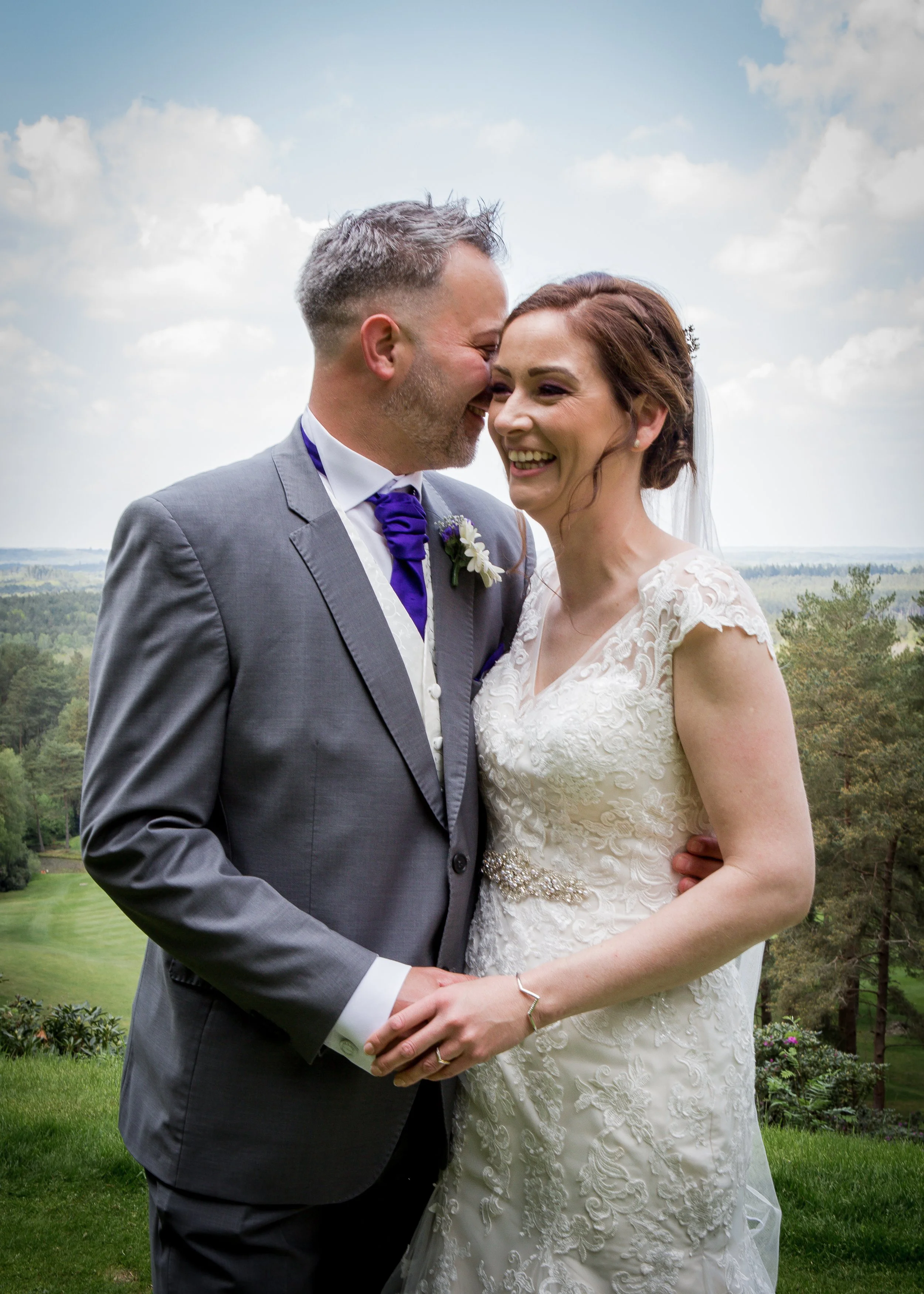 A newlywed couple is standing outdoors on a lush green landscape with trees and cloudy sky in the background. The groom is wearing a gray suit with a purple tie and boutonniere, and the bride is in a white lace wedding dress with a train and veil. Th