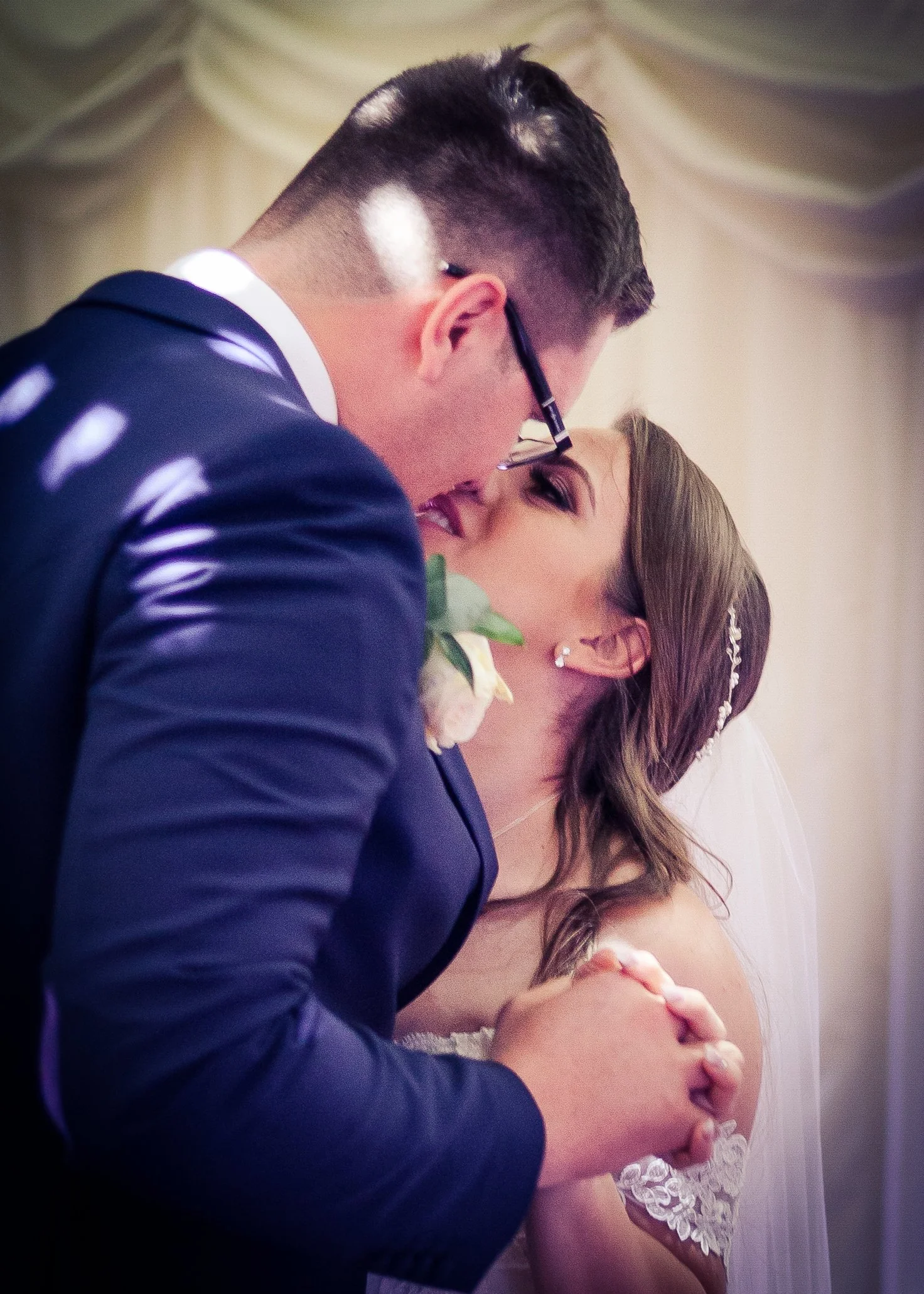 A groom in a navy suit leaning over and kissing his bride on her forehead, who is wearing a white lace dress and a tiara, during their wedding ceremony.